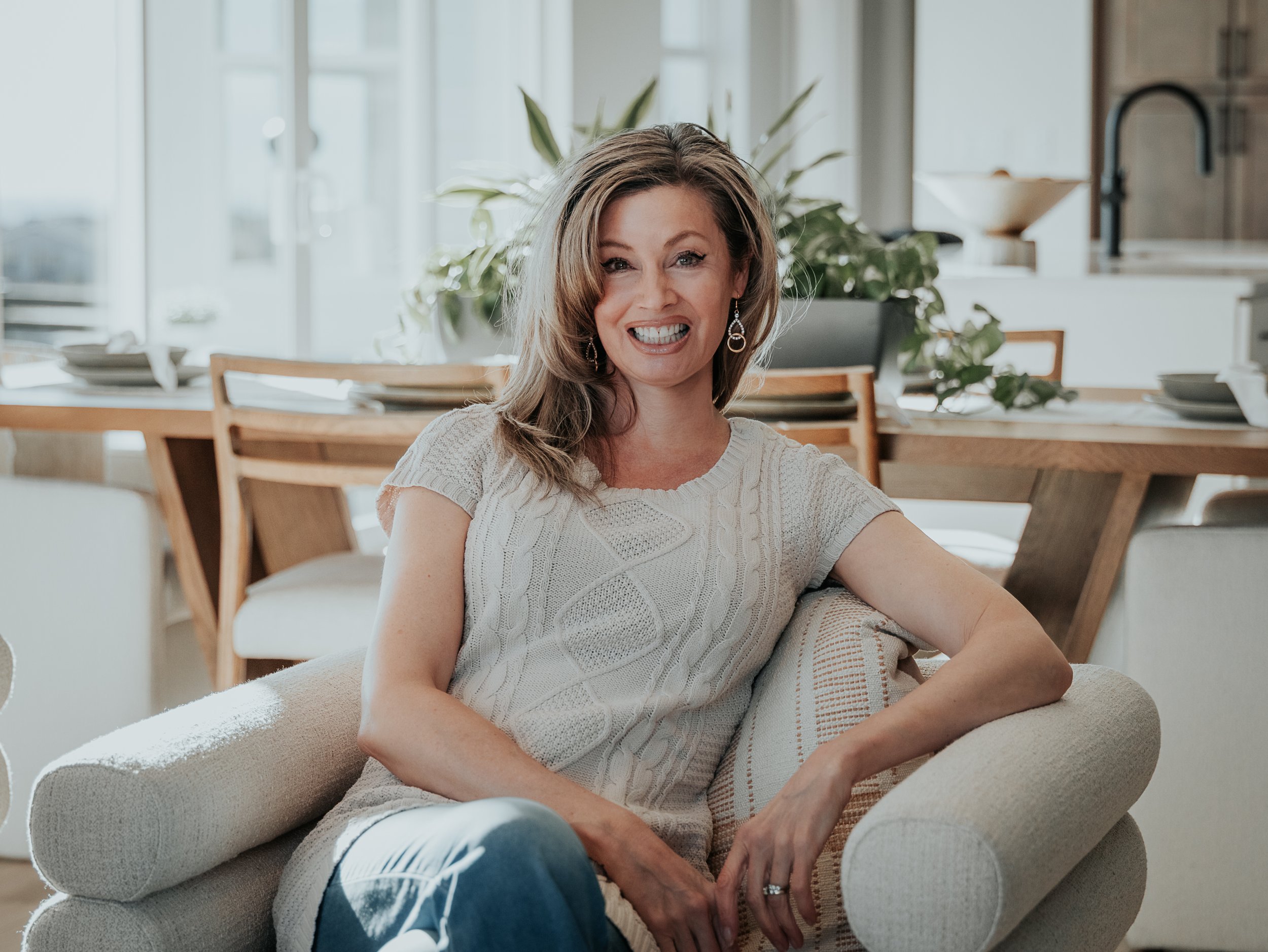 A woman with brown hair in a beige sweater sitting on a beige sofa in a well-lit room, smiling at the camera, with a dining area and green plants in the background.