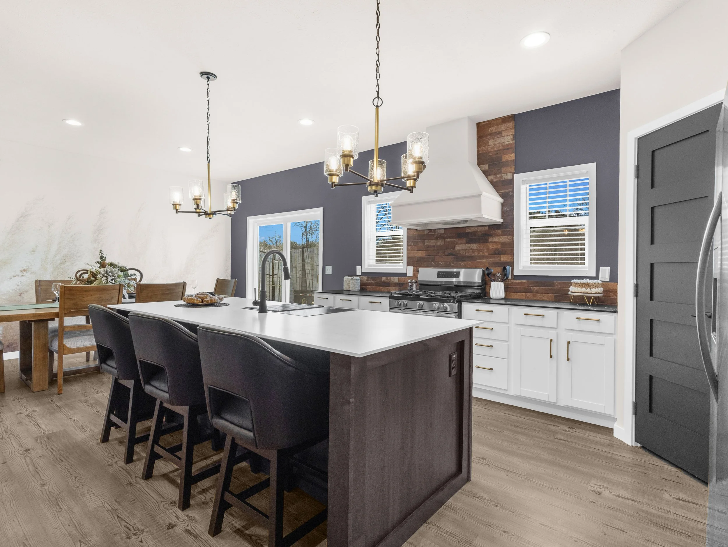 Open-concept kitchen with white island, black chairs, chandelier, brick accent wall, gray cabinets, and stainless steel appliances.