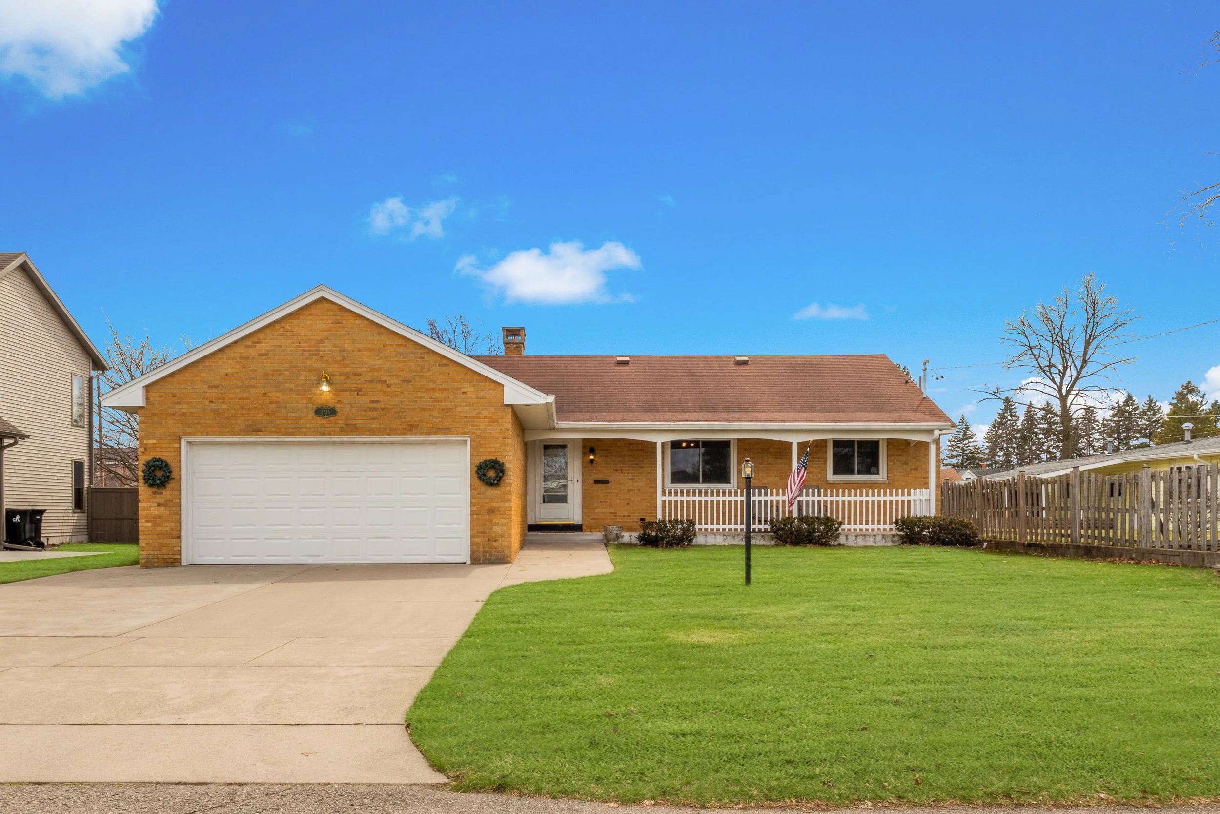 Front view of a suburban house with a brick facade, white garage door, porch with white railings, American flag, and a well-maintained lawn. Clear blue sky with a few clouds in the background.