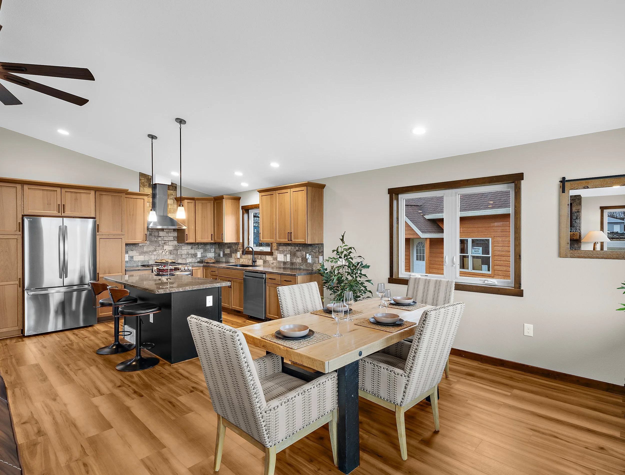 Interior view of a modern kitchen and dining area with wooden cabinets, stainless steel refrigerator, brick backsplash, and a wooden dining table surrounded by upholstered chairs. Large windows allow natural light into the space.