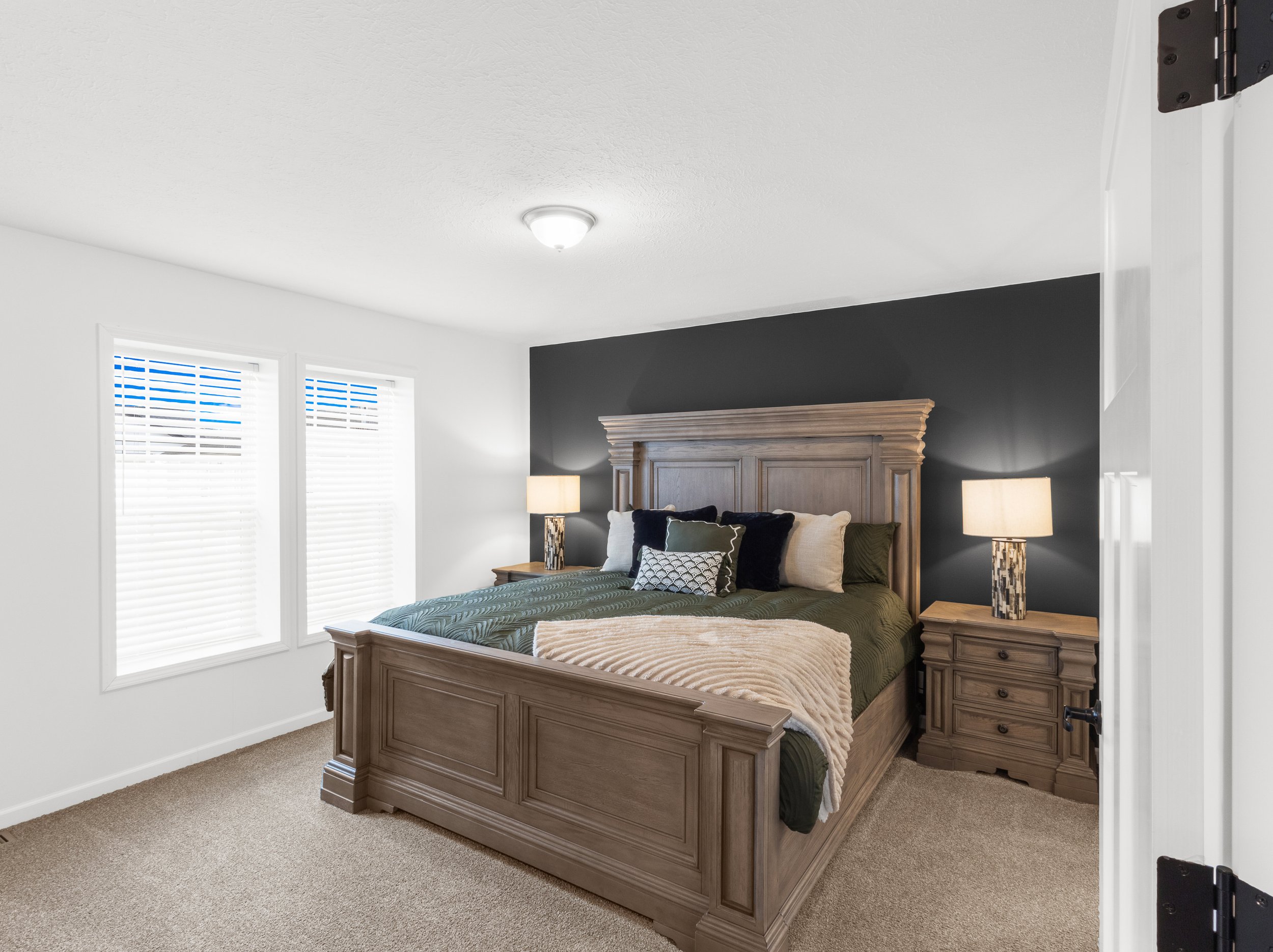 A bedroom with a wooden bed frame, dark bedding, and a beige blanket. Two nightstands with lamps on each side of the bed. Two windows with blinds, a dark accent wall, and a ceiling light.