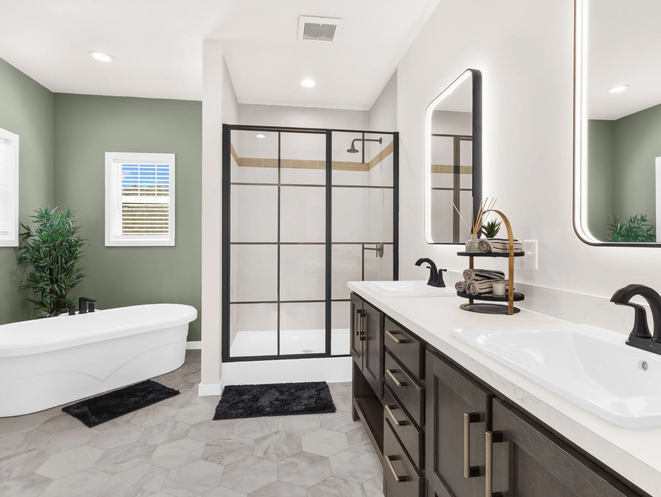 Modern bathroom with green accent wall, white bathtub, glass shower enclosure with black frame, double sink vanity with dark cabinets, oval mirrors with LED borders, and decorative plants.
