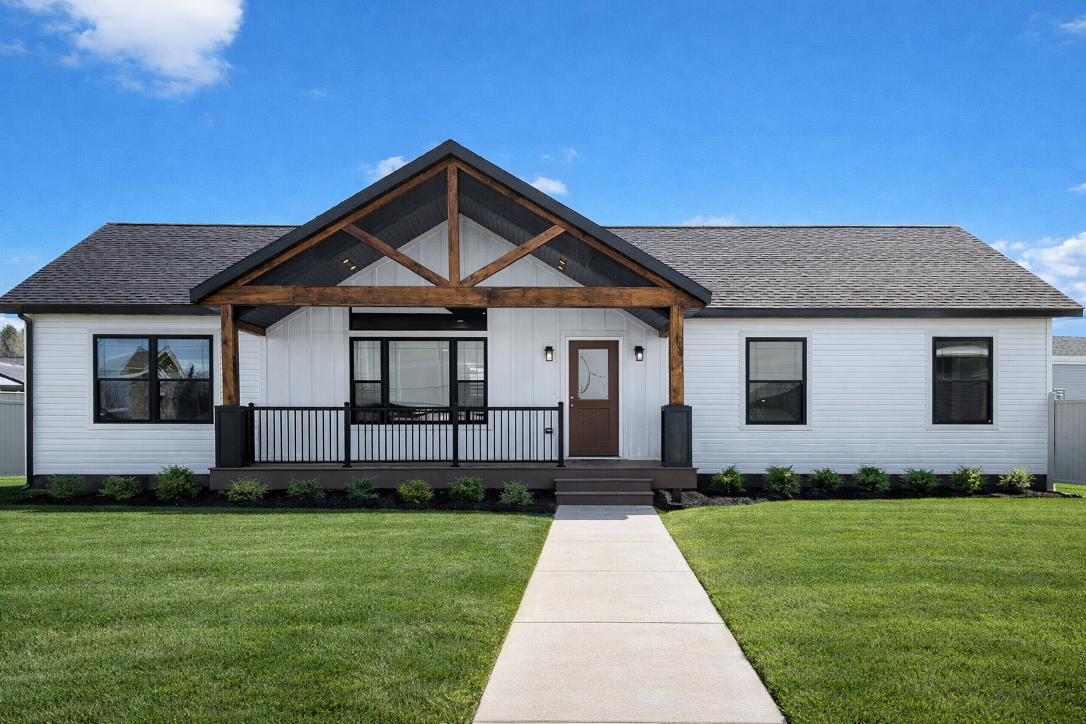 Modern single-story house with white siding, black-framed windows, a front porch with black railing, wooden beams overhead, and a concrete walkway leading to the front steps, surrounded by green lawn and small bushes, under a blue sky.