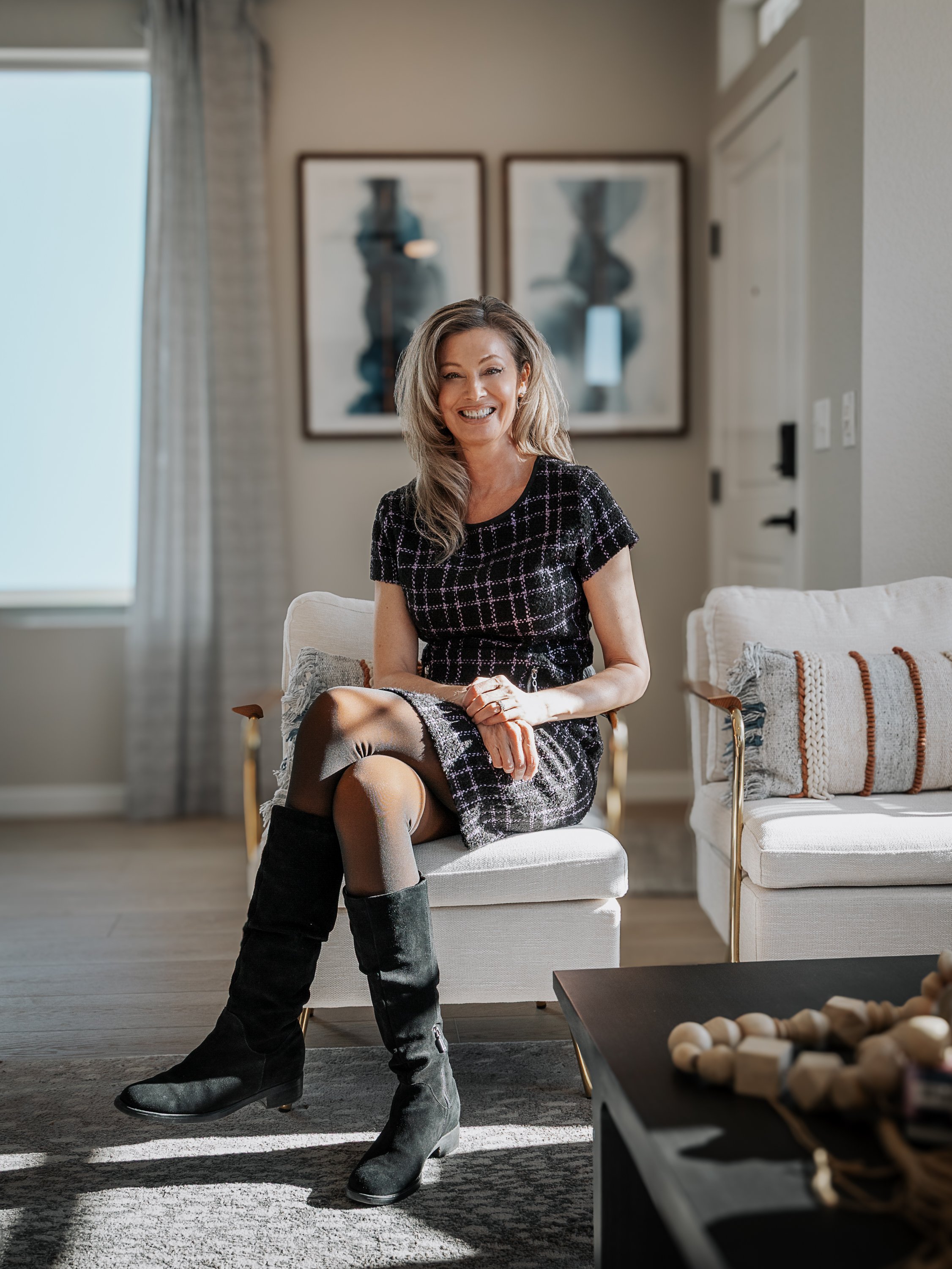 A woman with blonde hair smiling and sitting on a beige upholstered chair in a modern living room. She is wearing a black and white checkered dress with black knee-high boots. The room has natural lighting, a sliding glass door, and two framed abstra