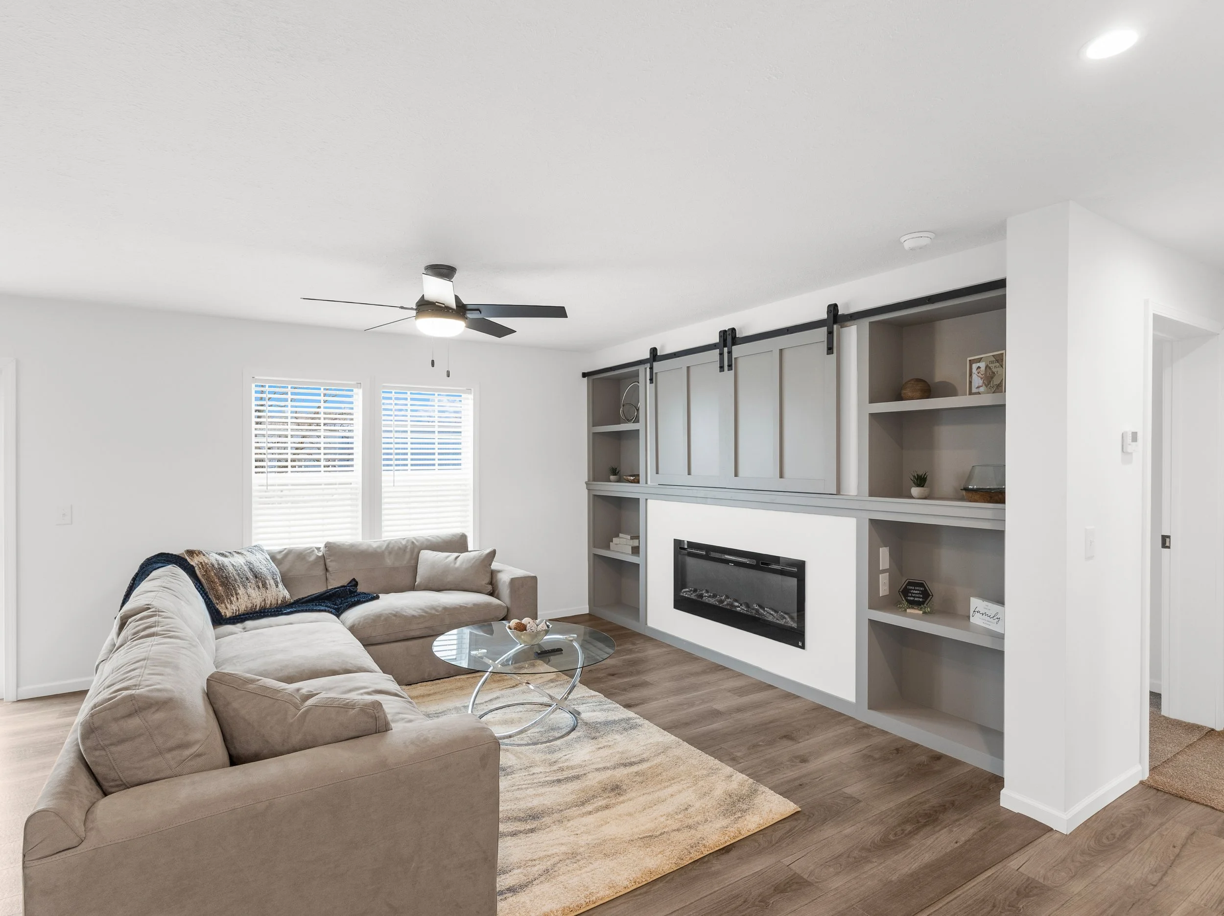 A modern living room with a beige sectional sofa, a glass coffee table, a built-in fireplace, and cabinetry with decorative items. There are two windows with white blinds and a ceiling fan with a light. The room has wooden flooring and neutral-colore