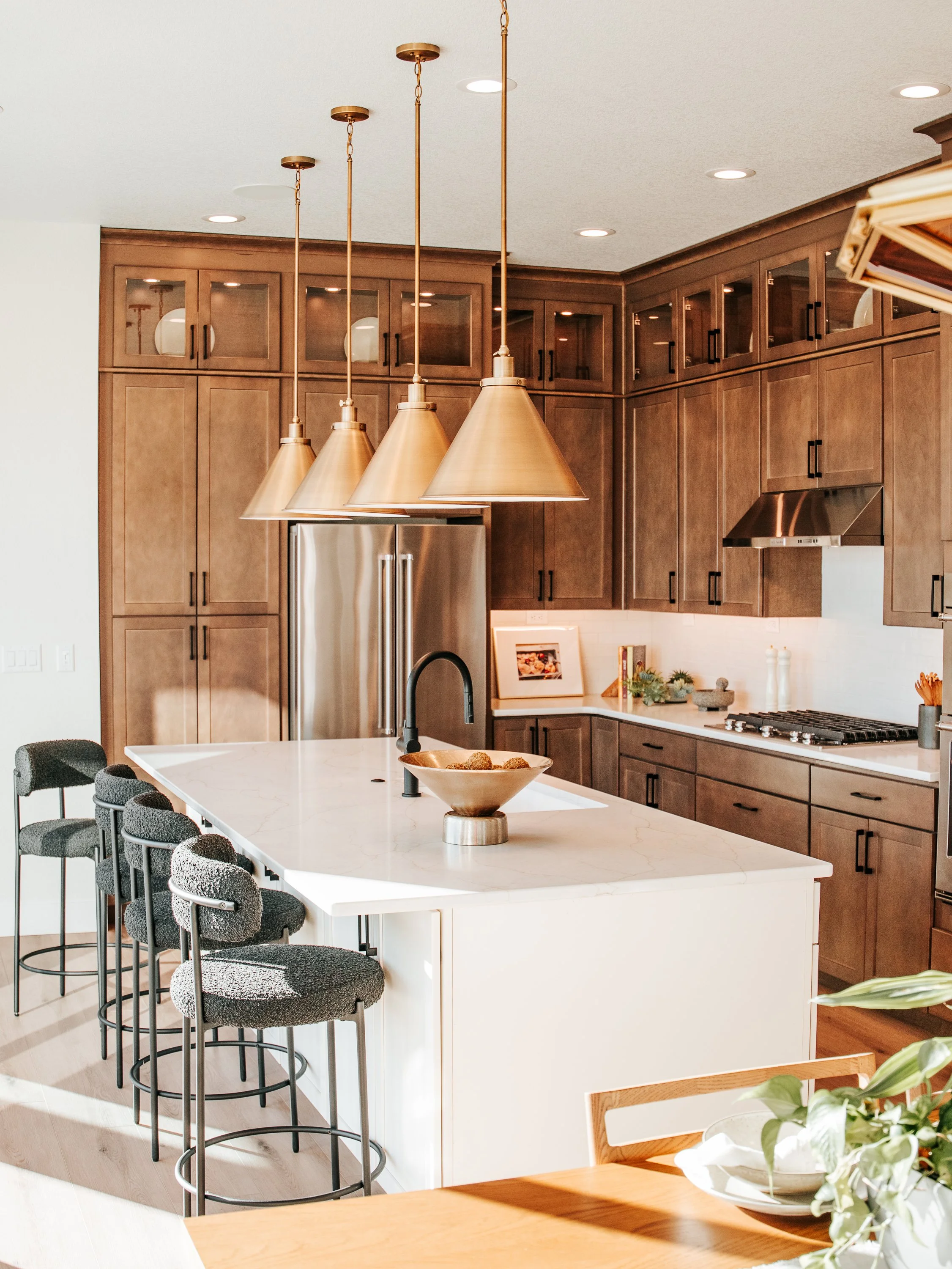 Modern kitchen with walnut cabinets, stainless steel appliances, and a large white marble island counter. Four gold pendant lights hang above the island. Black dining chairs surround a wooden dining table with a white bowl and green plants.