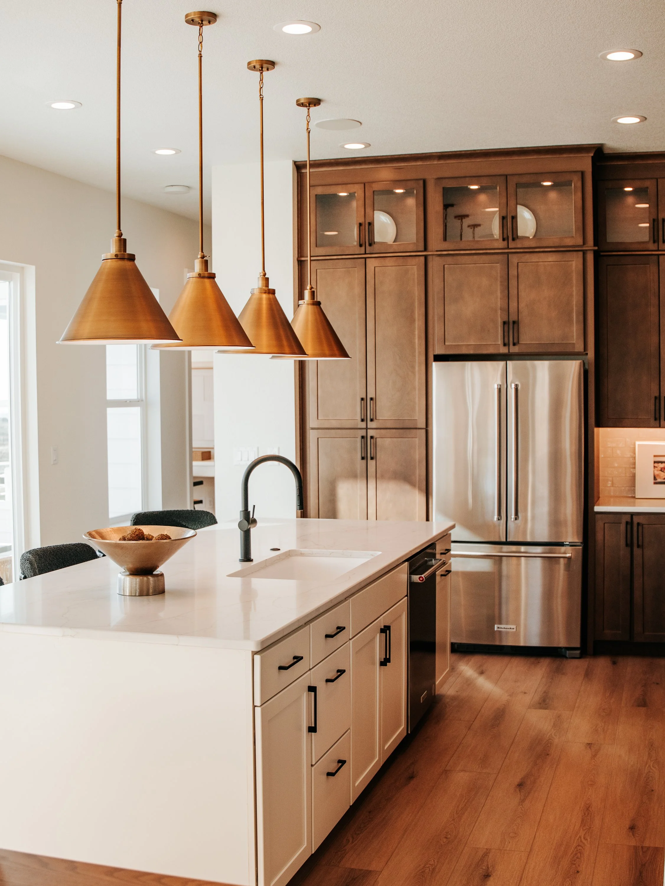 Modern kitchen with white island, copper pendant lights, wooden cabinets, stainless steel refrigerator, and hardwood floors.