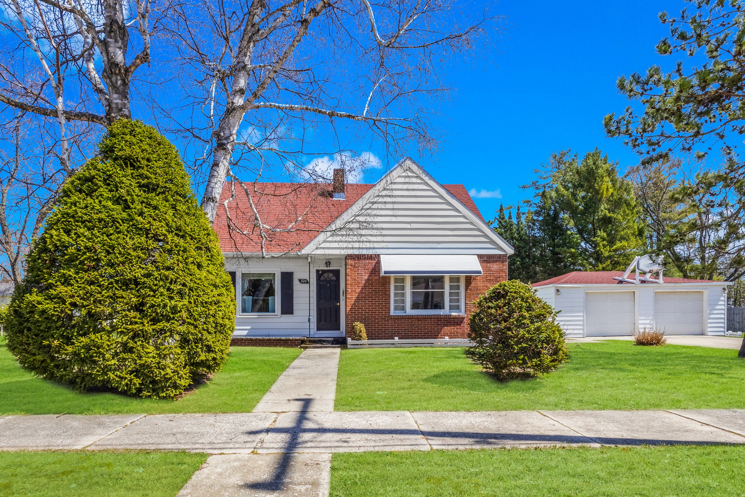 Front view of a house with a brick and white siding exterior, a red tile roof, a chimney, large front window, door with a decorative screen, surrounded by green lawn, bushes, and trees, with a driveway and garage to the right.