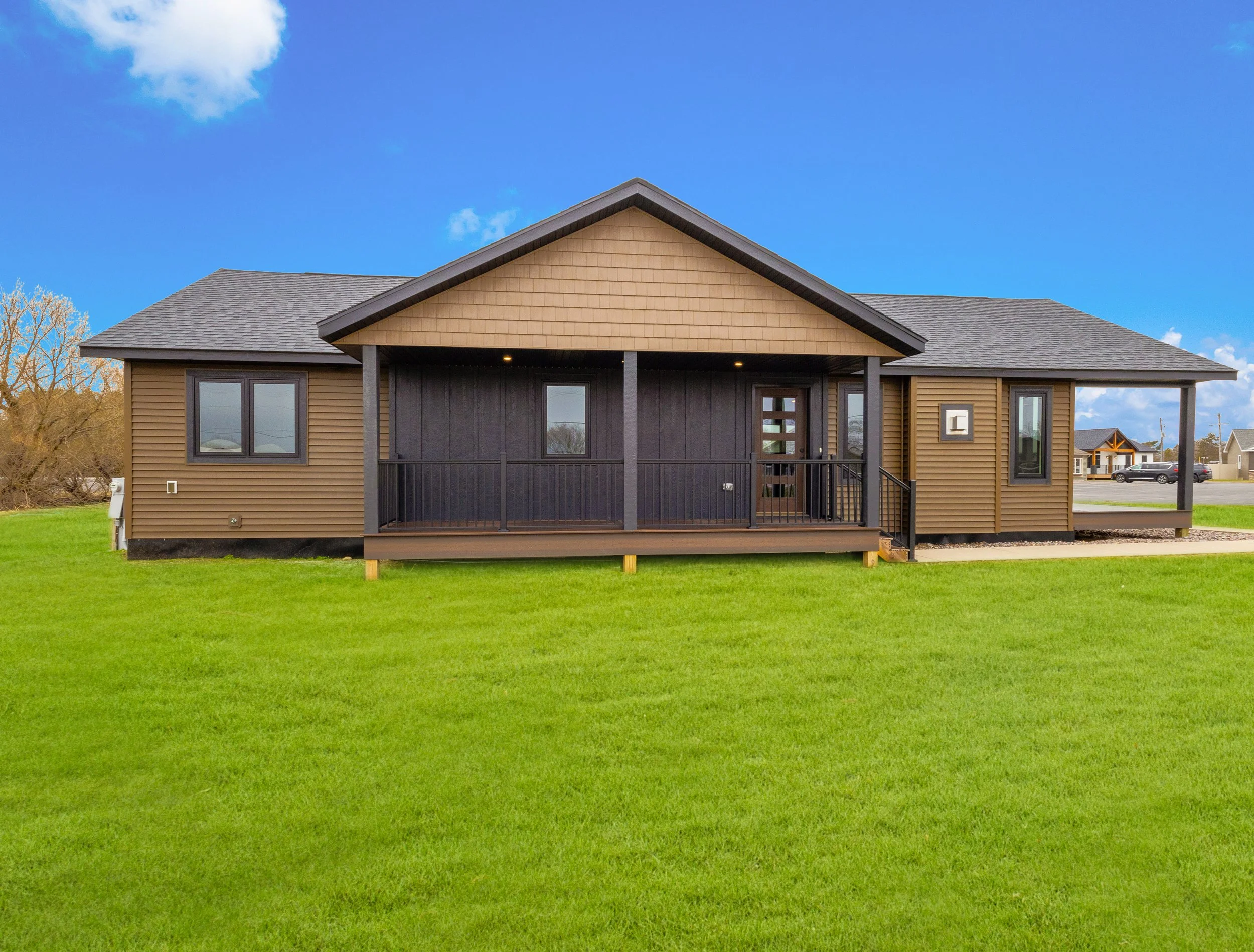 A modern house with tan and black exterior, front porch, and a green lawn under a blue sky with some clouds.