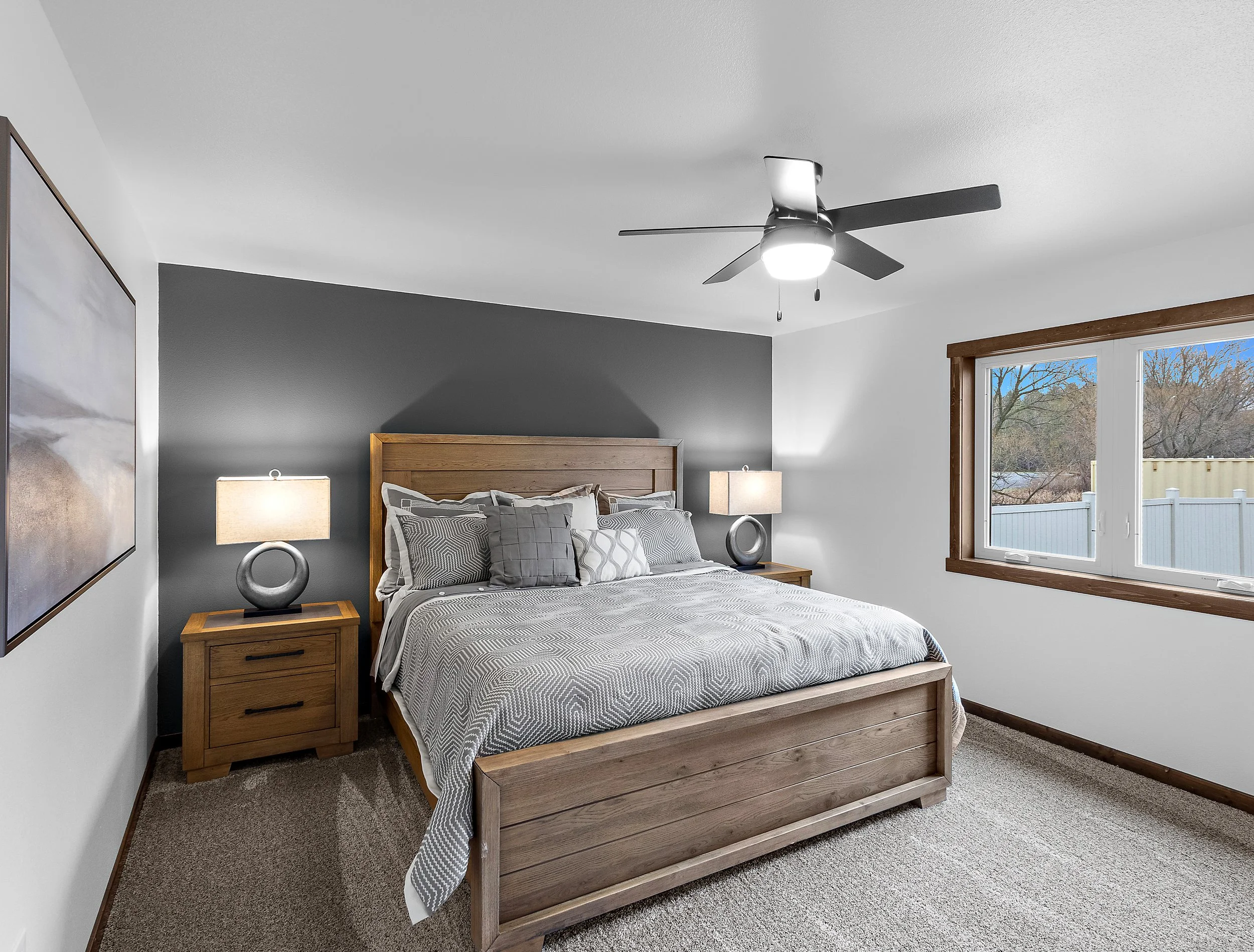 A bedroom with a wooden bed frame, gray and white bedding, two matching bedside tables with lamps, a window with brown trim, and a ceiling fan with lights.