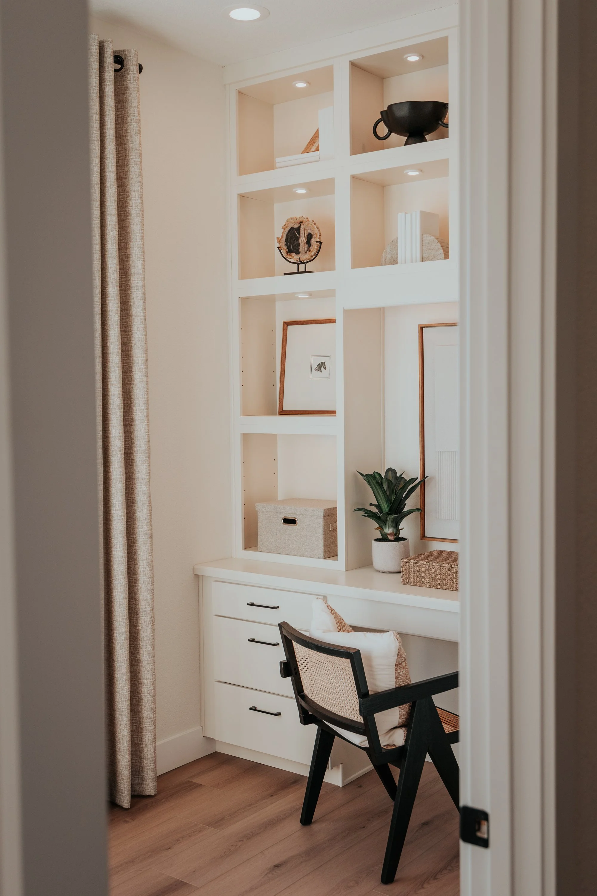 A small home office nook with built-in white shelving and drawers, a black chair with a beige cushion, a potted plant, framed artwork, decorative objects, and beige curtains.