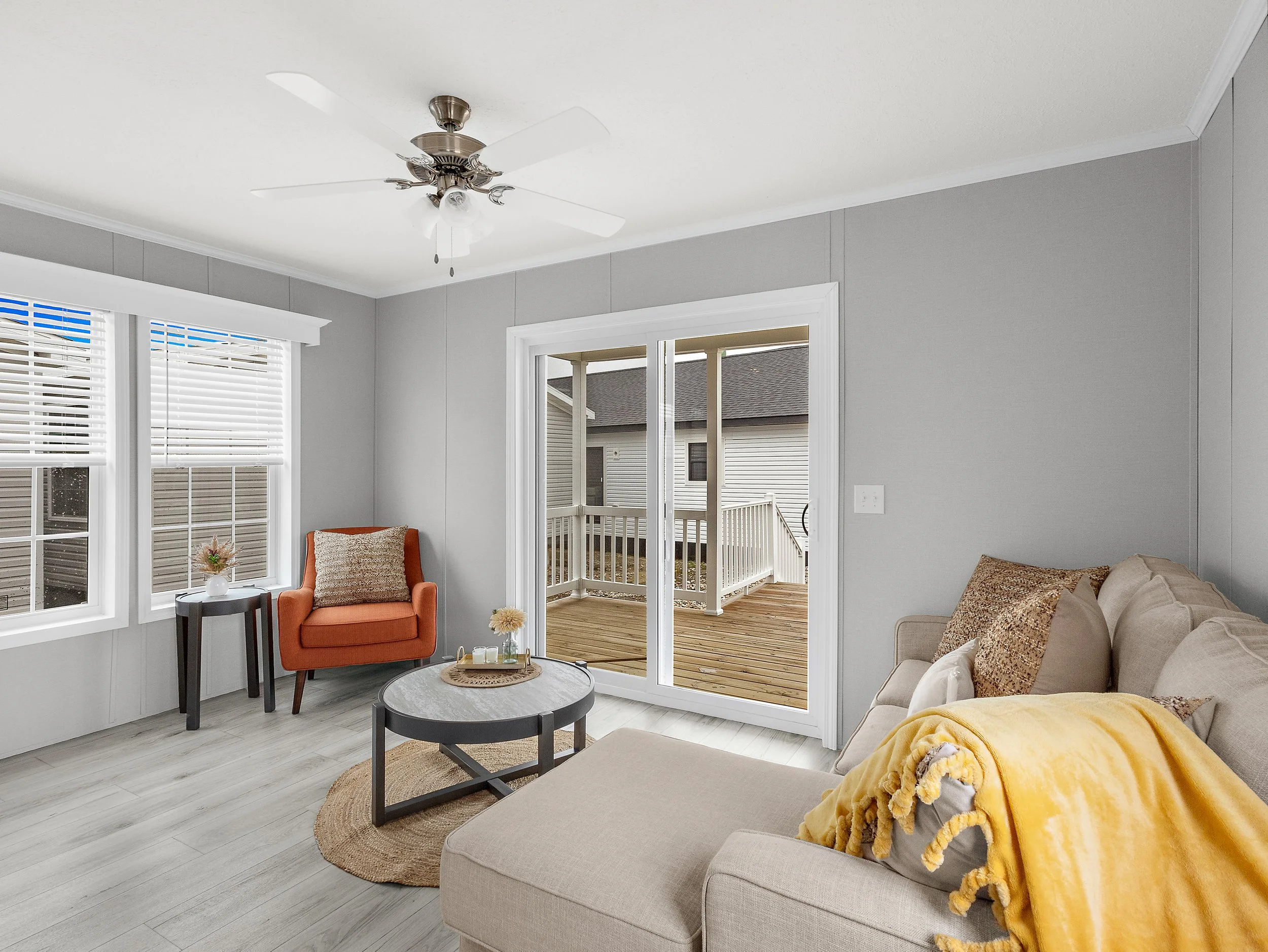 Living room with light gray walls, large windows with white blinds, sliding glass door leading to a deck, beige sofa with throw pillows, orange armchair, small round coffee table, side table, and ceiling fan.
