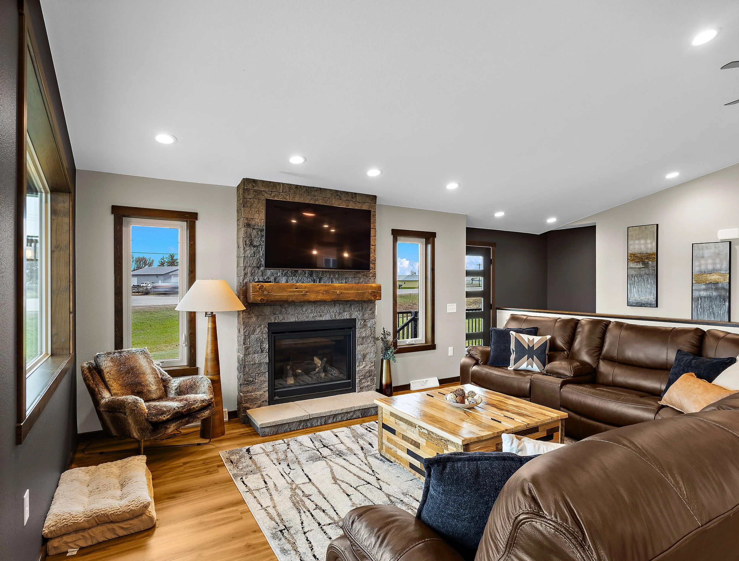 Living room with a stone fireplace, mounted television, leather sofa, colorful pillows, wooden coffee table, armchair with fur throw, and windows showing an outdoor view.