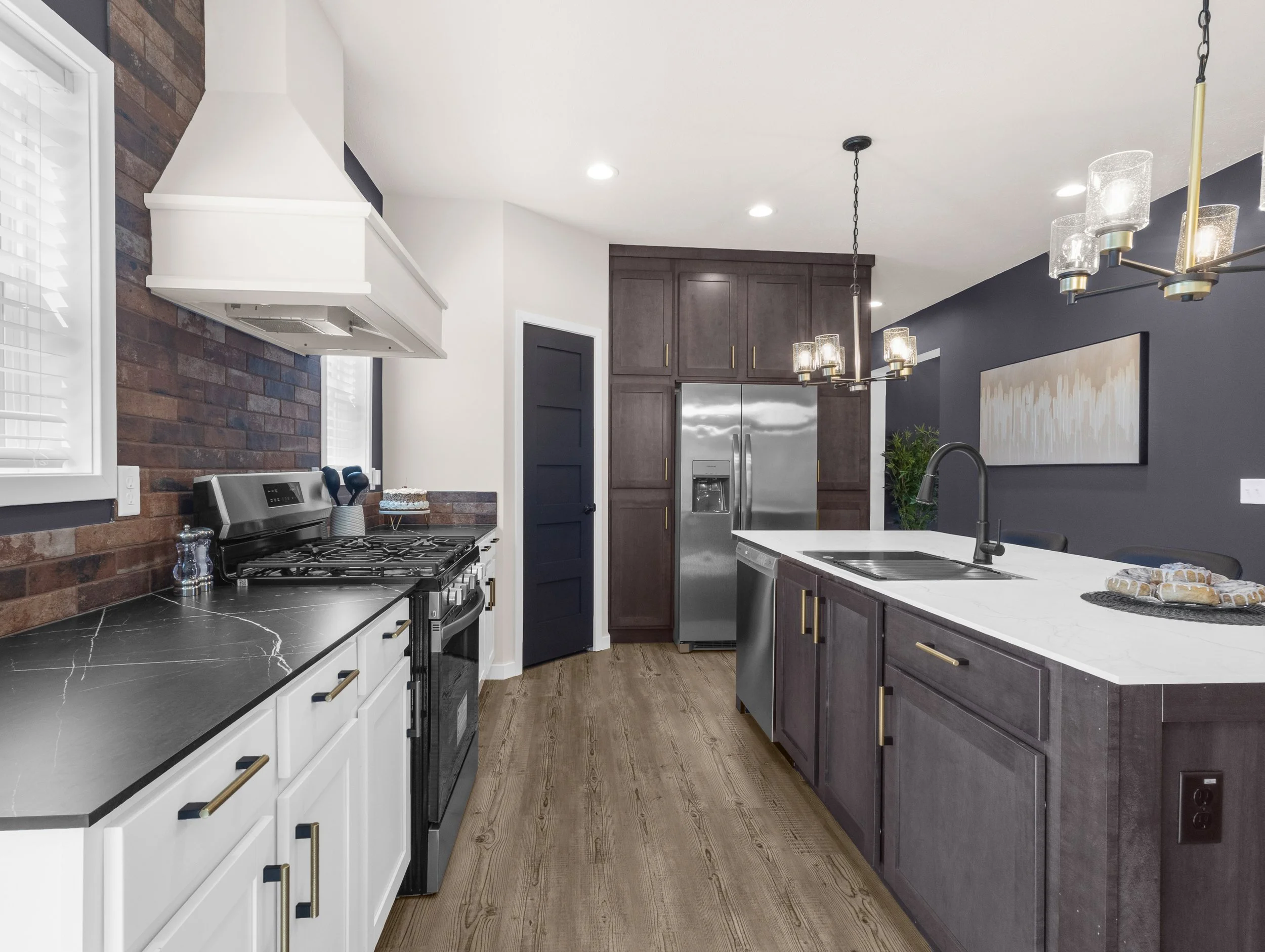 Modern kitchen with dark purple cabinets, white countertops, stainless steel appliances, and a brick accent wall. There are white kitchen cabinets on the left, a stainless steel refrigerator, and a black sink with a black faucet on the right. Two pen