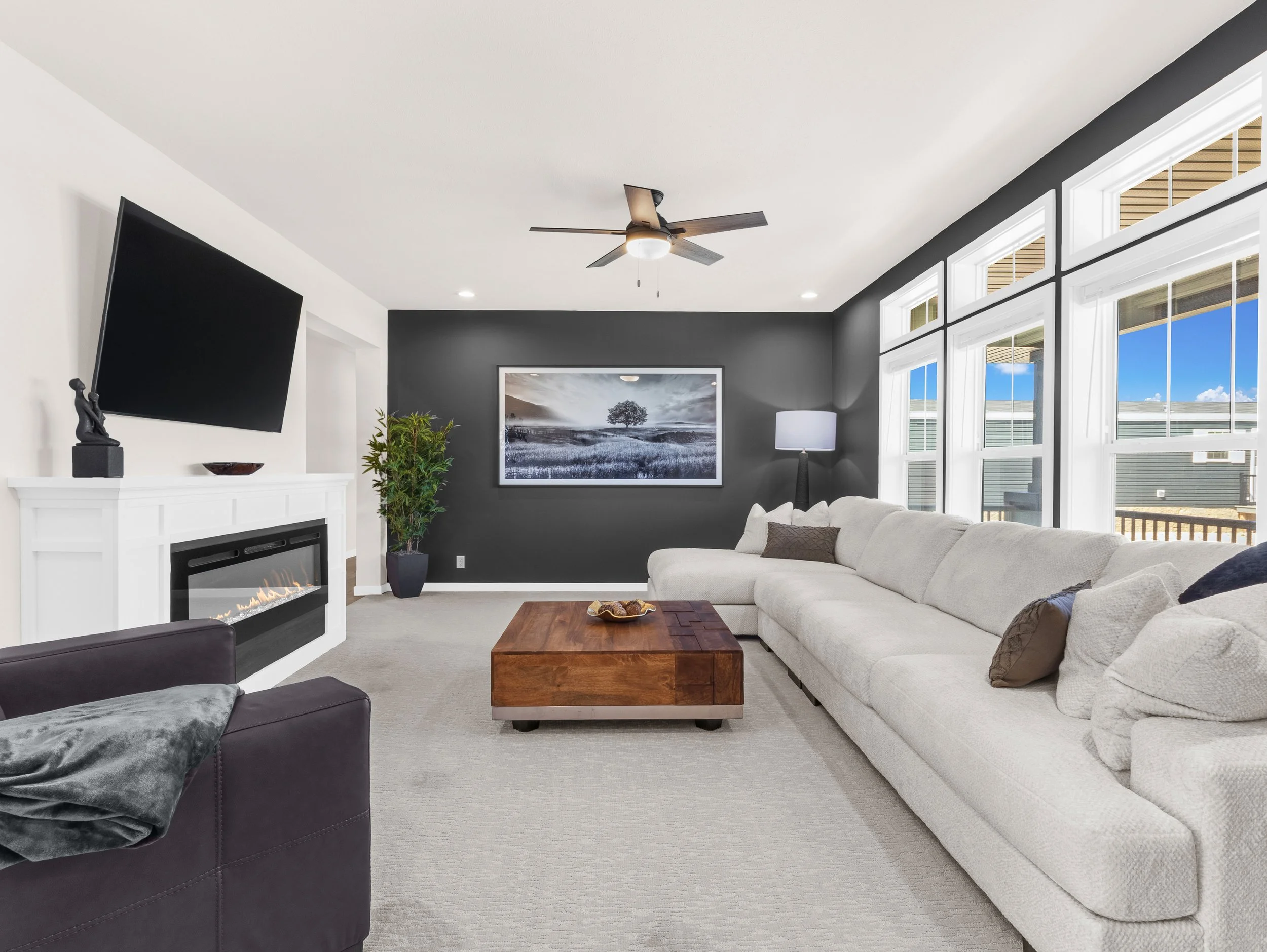 Living room with a white sectional sofa, dark gray accent wall, large windows, a wall-mounted TV, fireplace, ceiling fan, and artwork.
