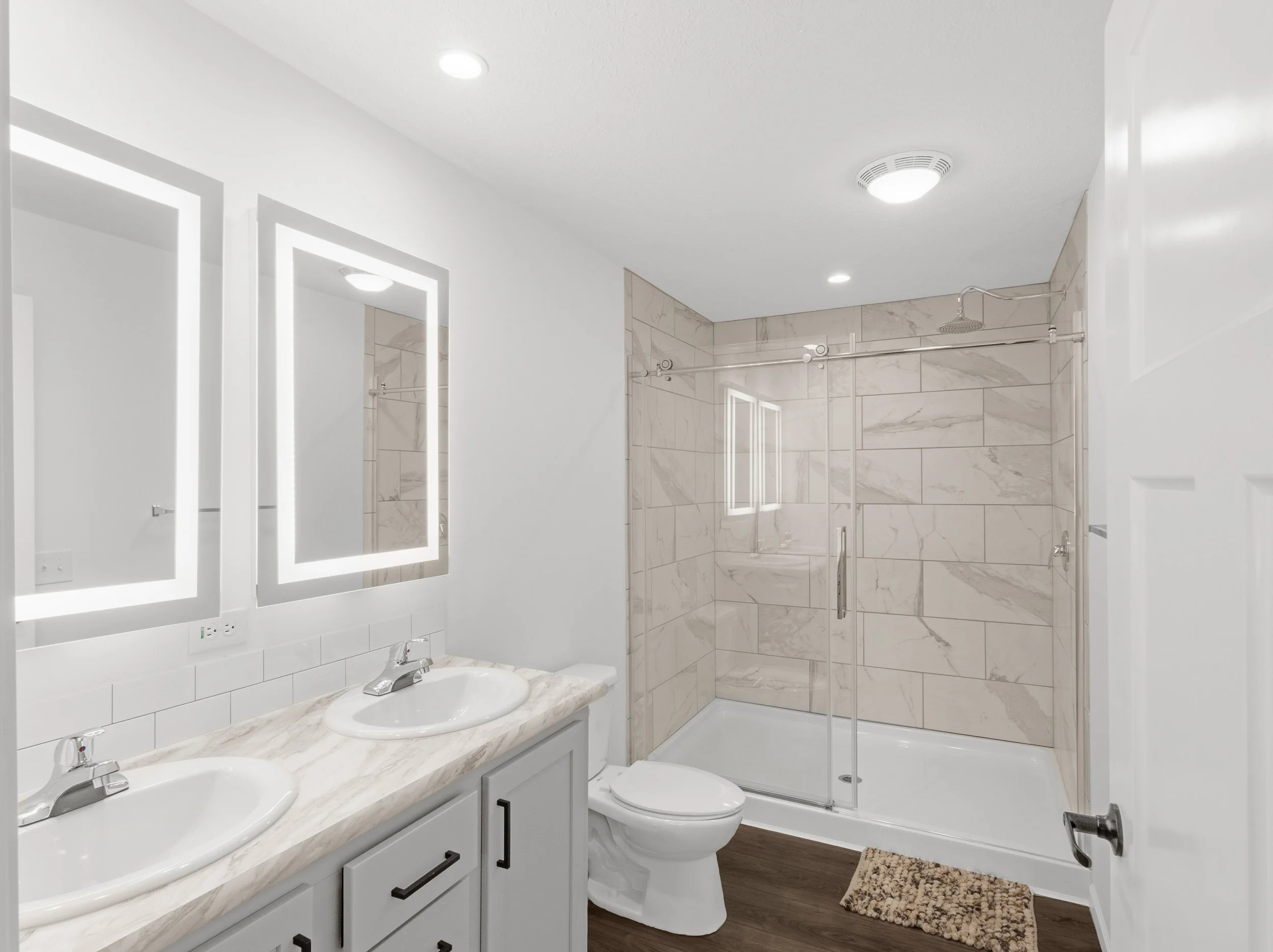 Modern bathroom with double sinks, large mirrors with LED lights, a toilet, and a walk-in shower with glass door, marble tiles, and ceiling lights.