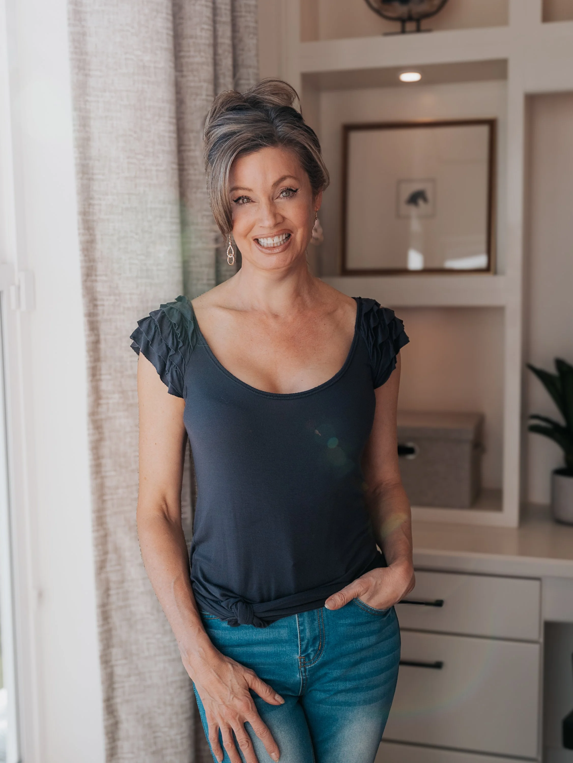 A woman with short gray hair wearing a black top and blue jeans, standing indoors near a doorway, smiling at the camera.
