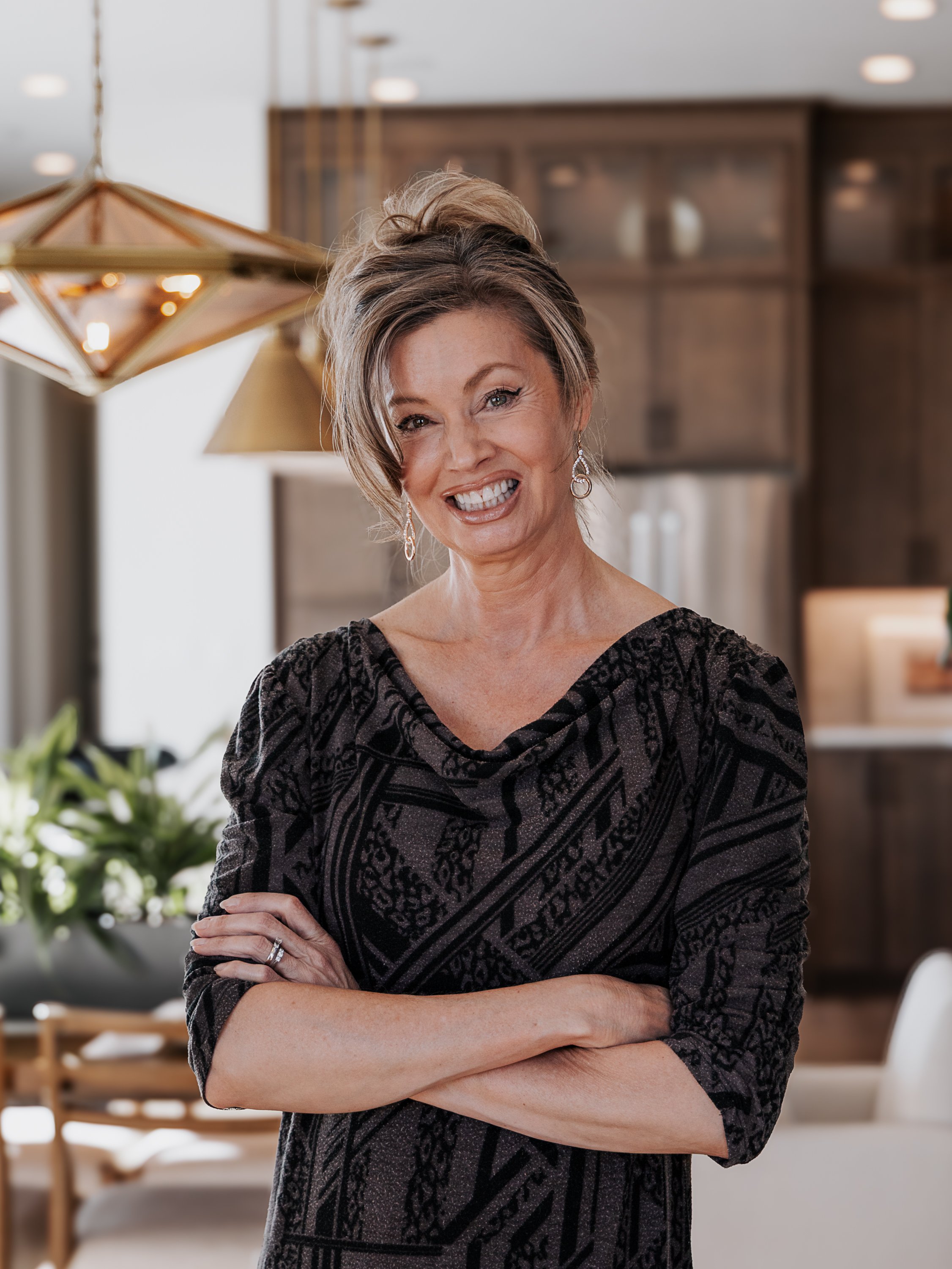 A woman with short brown hair, wearing earrings and a black patterned dress, smiling with arms crossed in a modern interior space with warm lighting and wooden furniture.