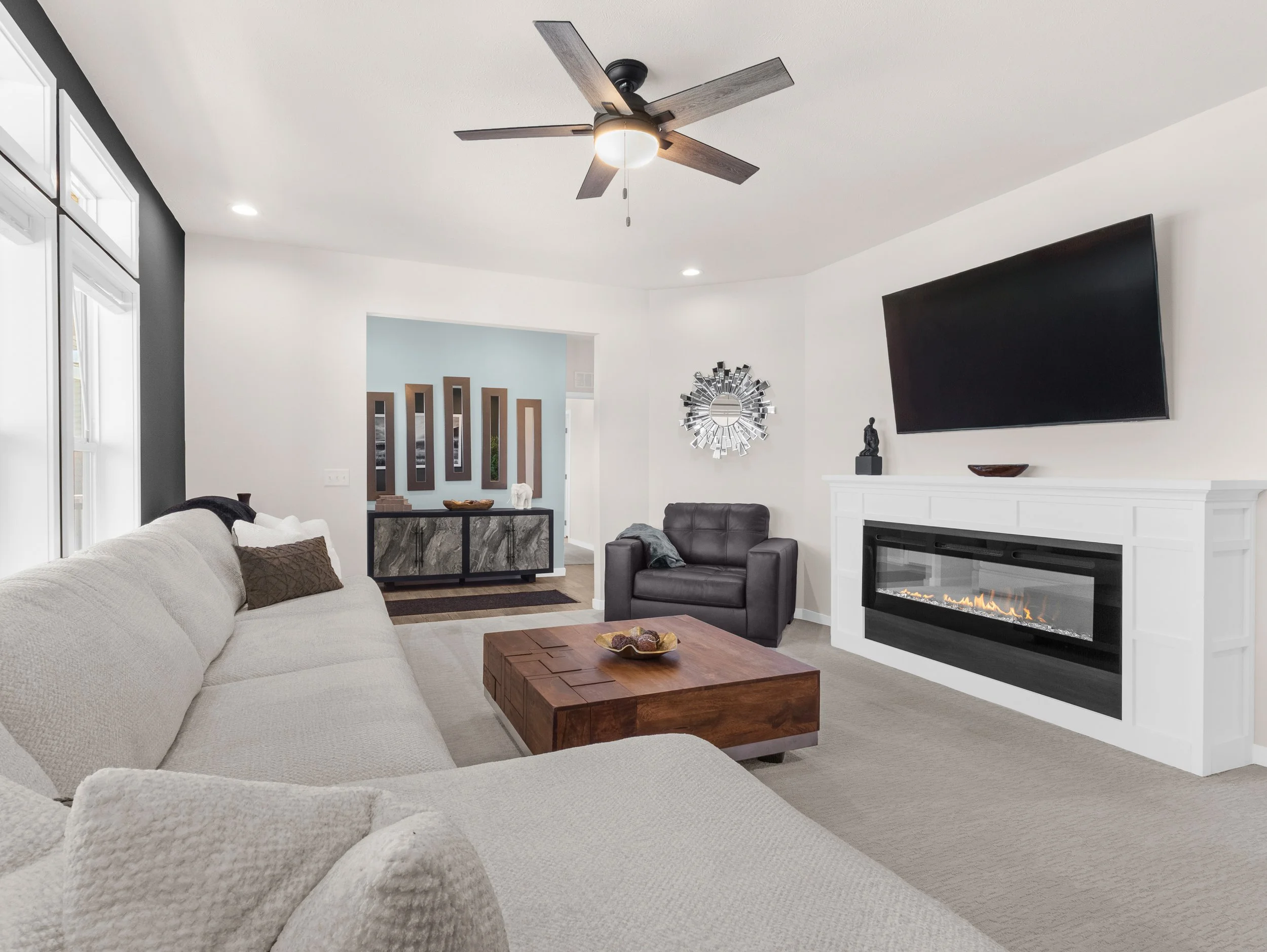 Modern living room with a large cream sofa, dark brown armchair, wooden coffee table, wall-mounted flat-screen TV, and fireplace with a decorative mantelpiece. Contains wall art, a sculpture, and a round mirror.