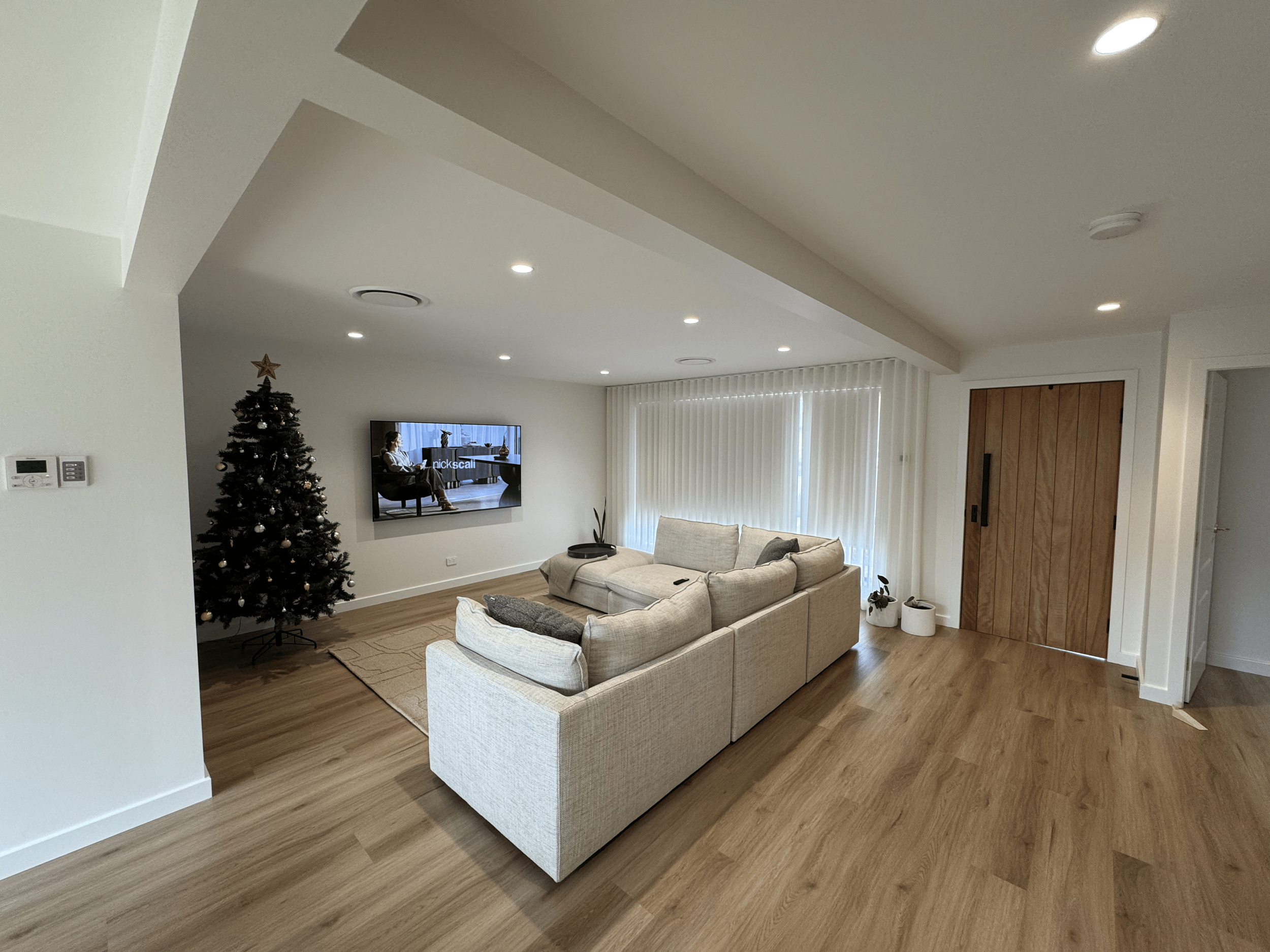 A living room with a Christmas tree, a beige sectional sofa, a wall-mounted television, wooden flooring, and sliding wooden door with vertical blinds covering the windows.