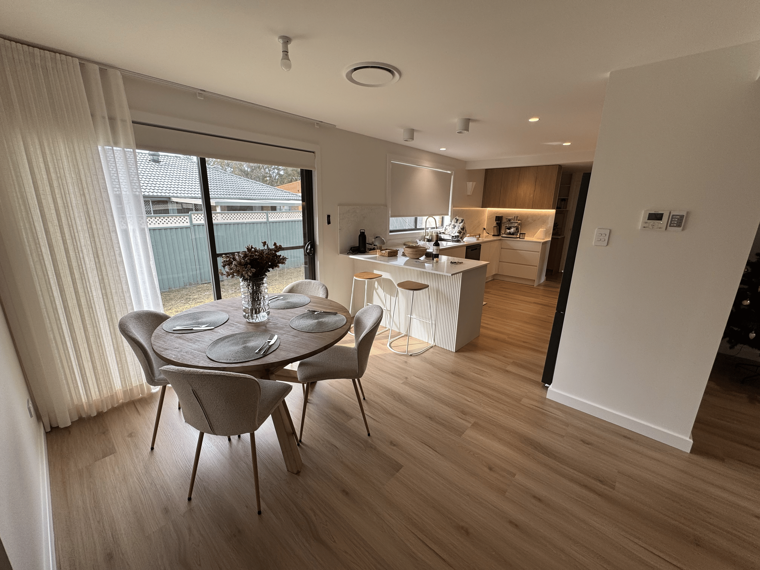 Open-concept dining and kitchen area with wooden flooring, round dining table with six chairs, vase with flowers, sliding glass door with curtains leading to outdoor yard, kitchen island with two stools, modern kitchen with beige cabinets, appliances, and counter with bowls and bottles.