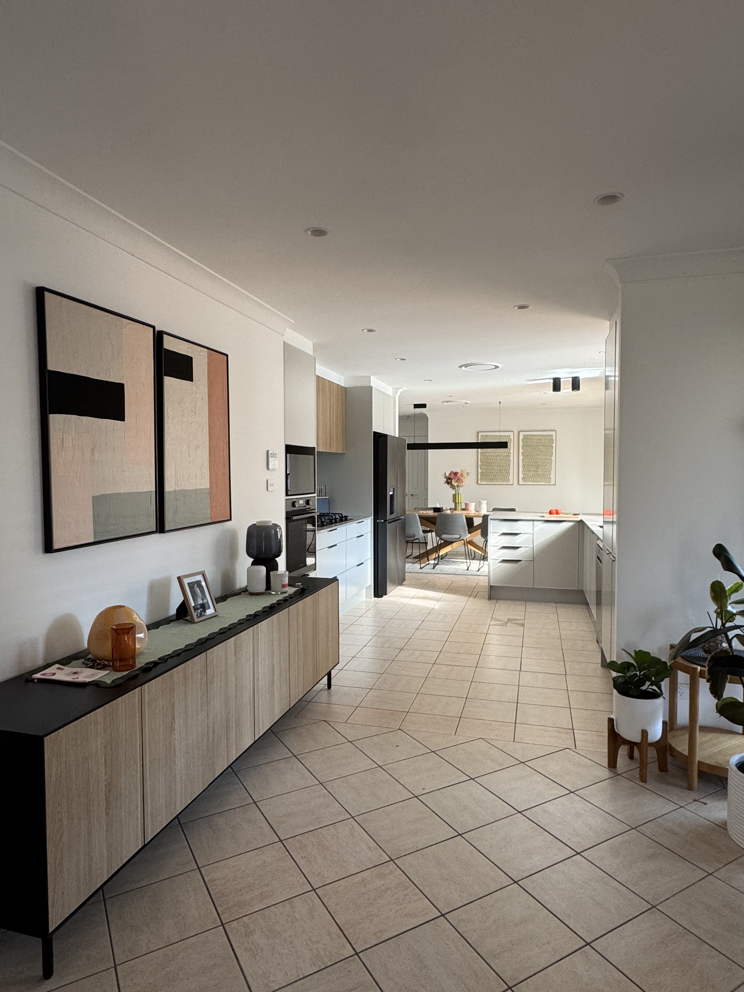 Modern open-concept kitchen and dining area with light-colored cabinets, black appliances, framed artwork, decorative vases, and potted plants.