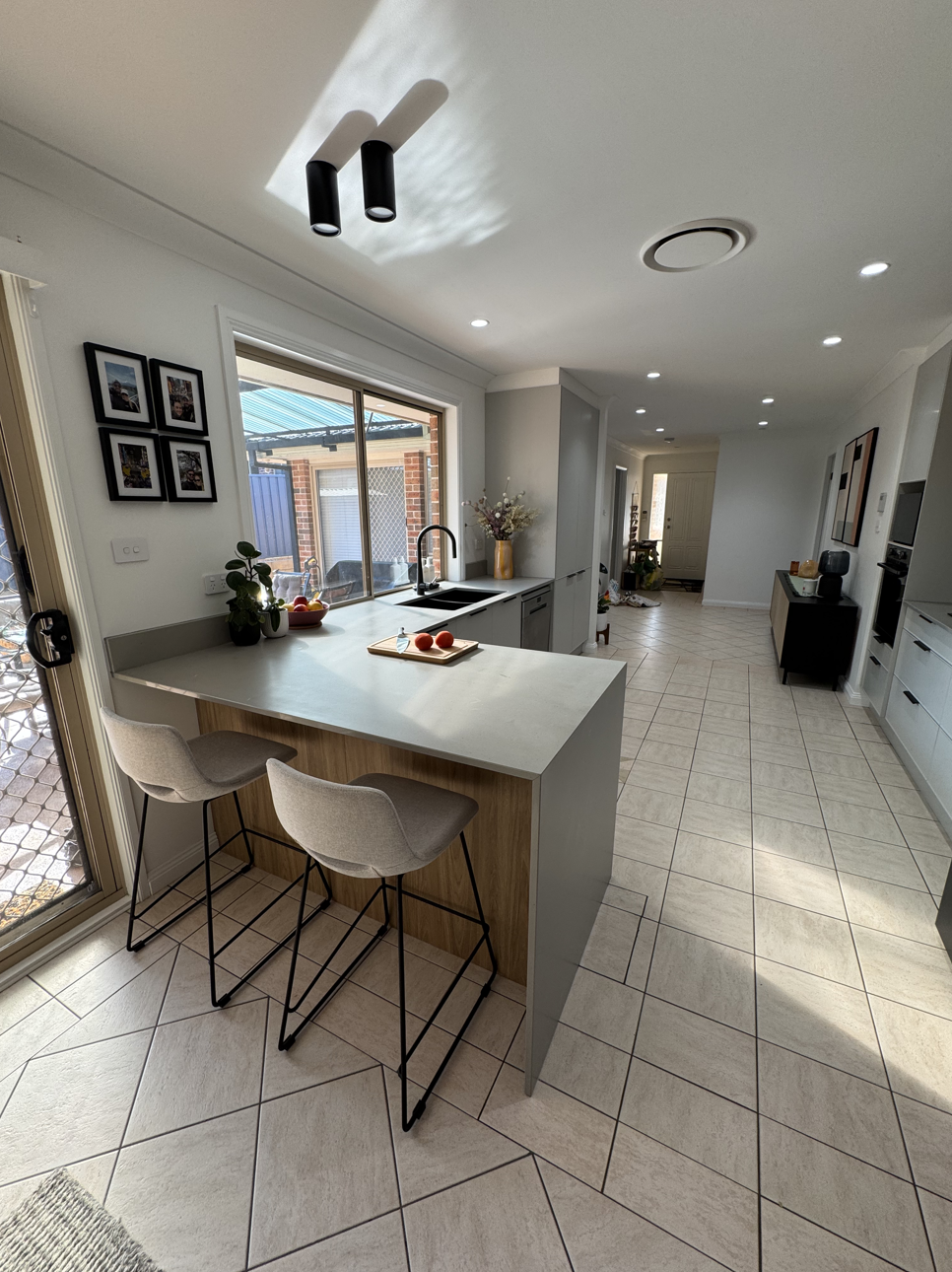 Modern kitchen with white countertops, two bar stools, a sink by a window, and home decor items.