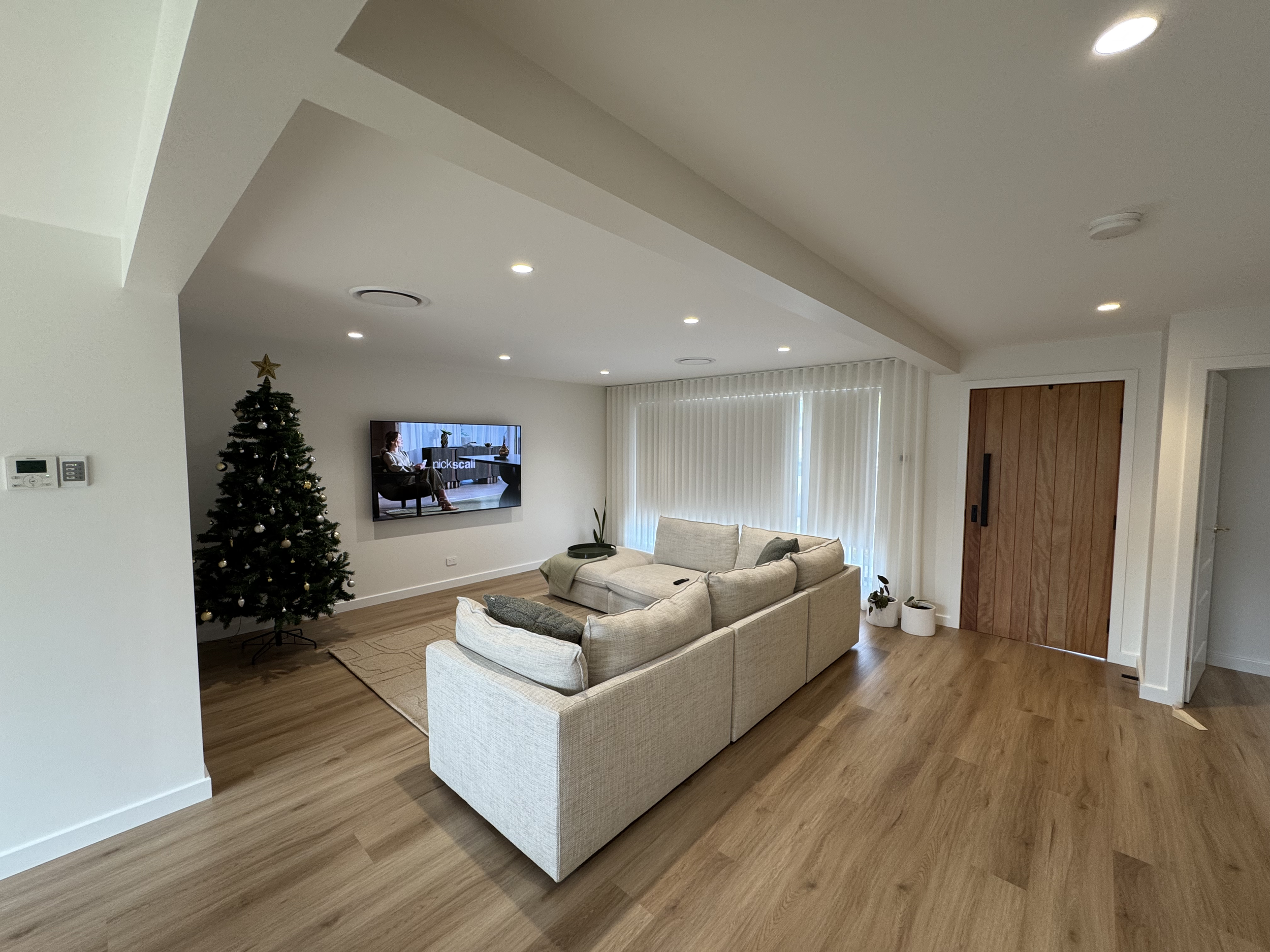 Living room with a beige sectional sofa, a wall-mounted television, a Christmas tree with ornaments and a star on top, wooden floor, window with vertical blinds, and some potted plants near the door.