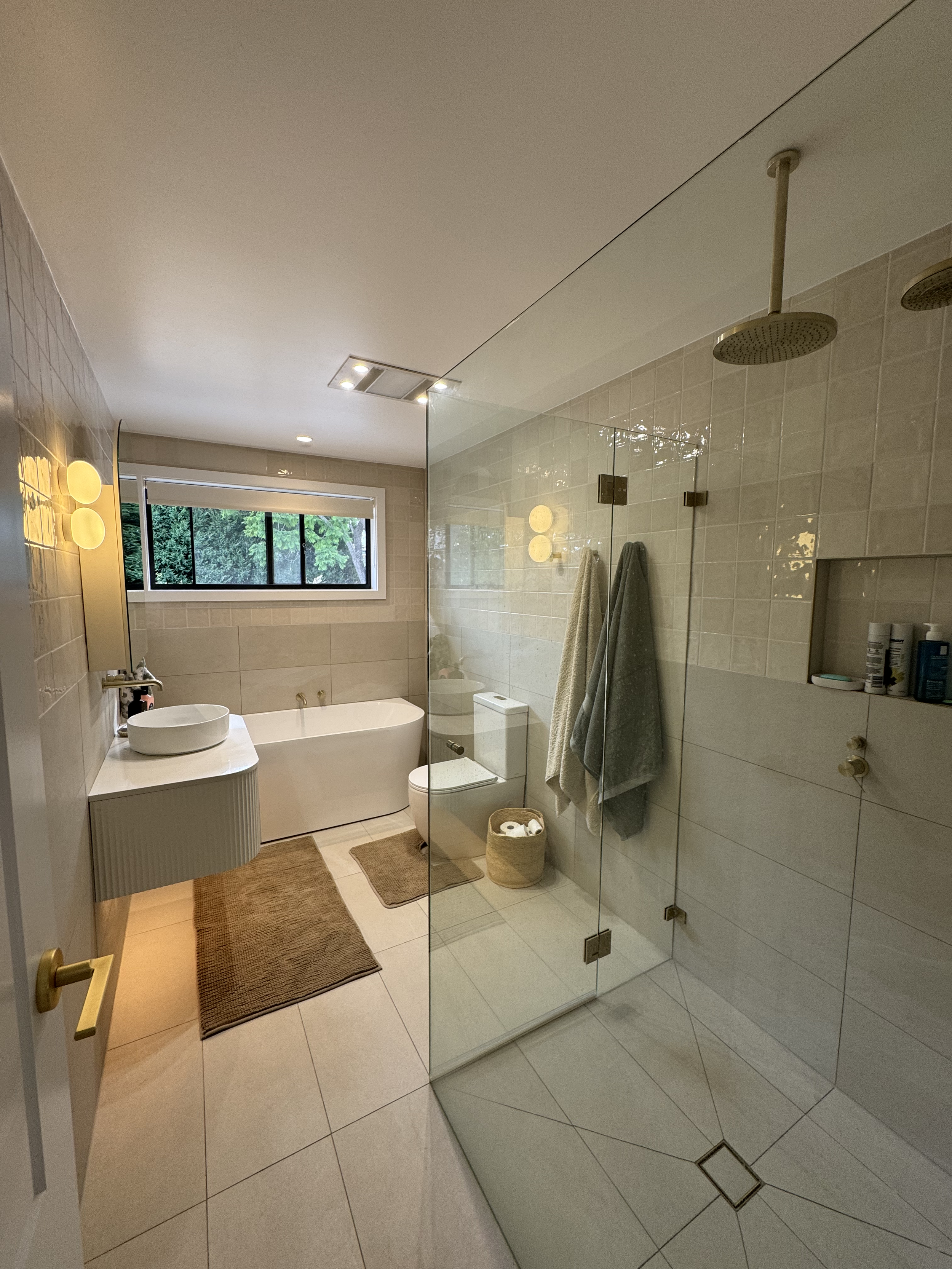 Modern bathroom with a bathtub, a white vanity with a vessel sink, a glass shower enclosure, a window with a view of greenery, towels hanging on hooks, and various toiletries on built-in shelves.