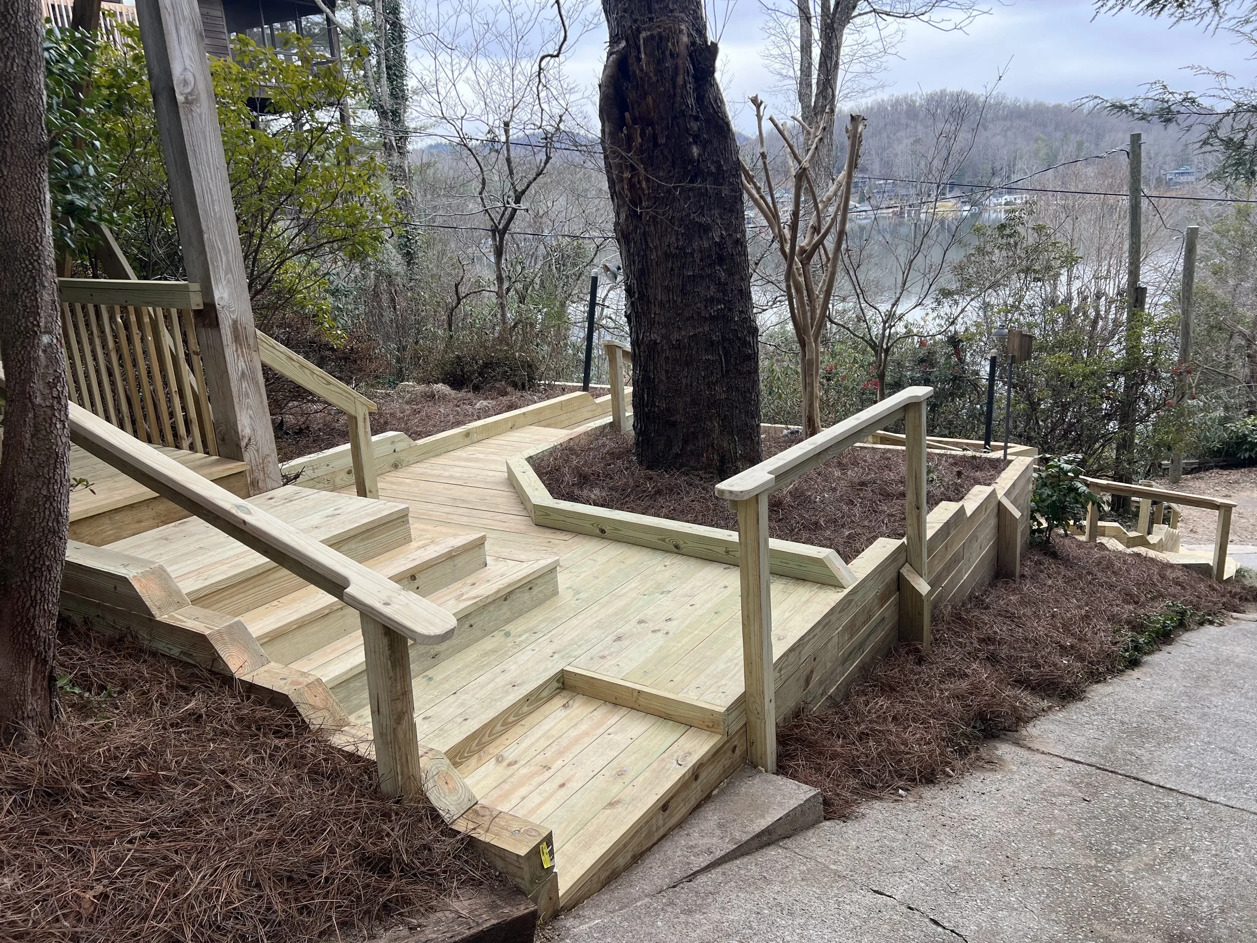 Newly constructed wooden outdoor staircase with handrails leading down to a landscaped yard with mulch and trees, overlooking a body of water in the background.
