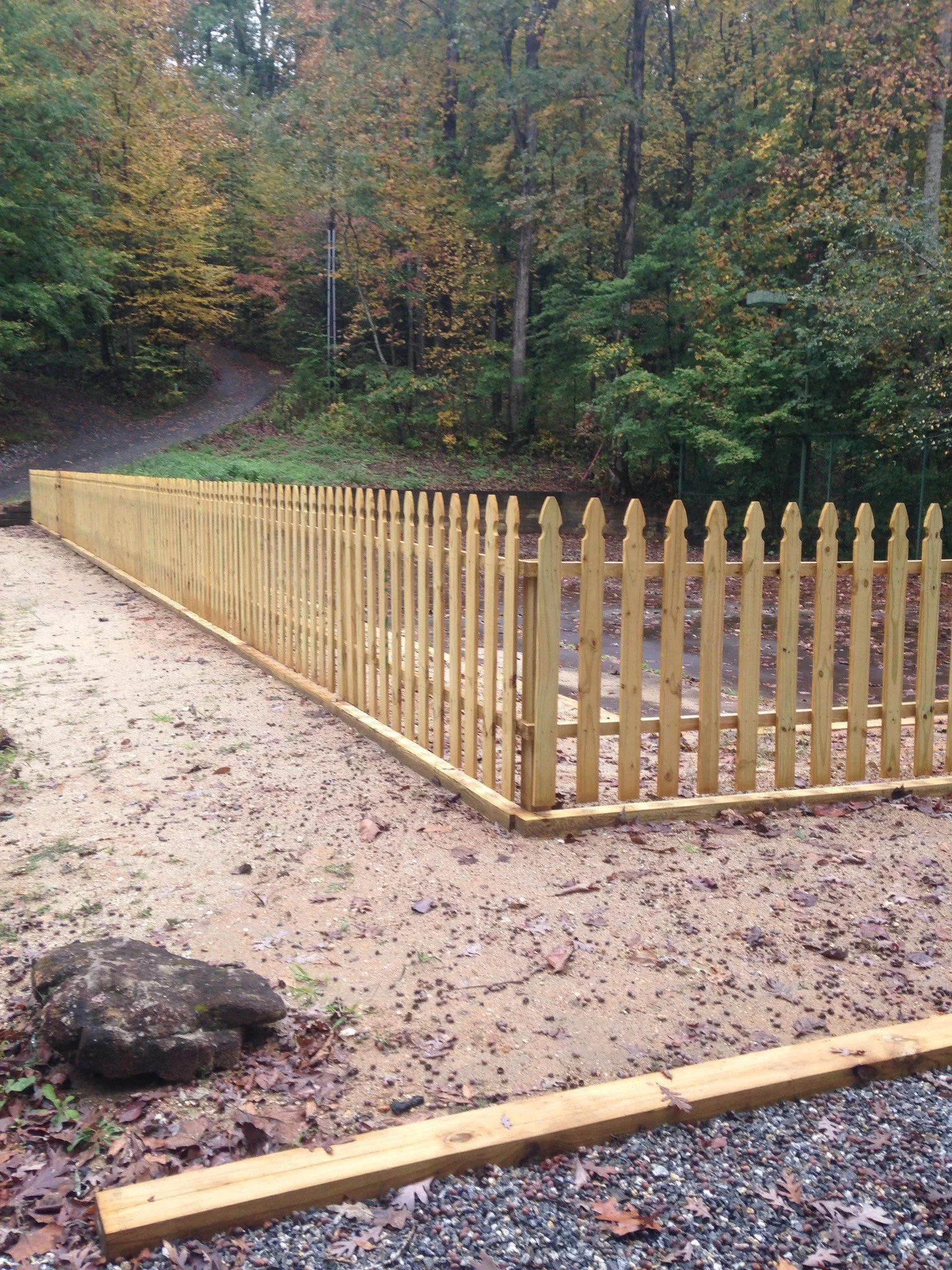 A newly installed wooden fence surrounds a small section of garden near a creek in a forested area, with a dirt path and autumn-colored trees in the background.