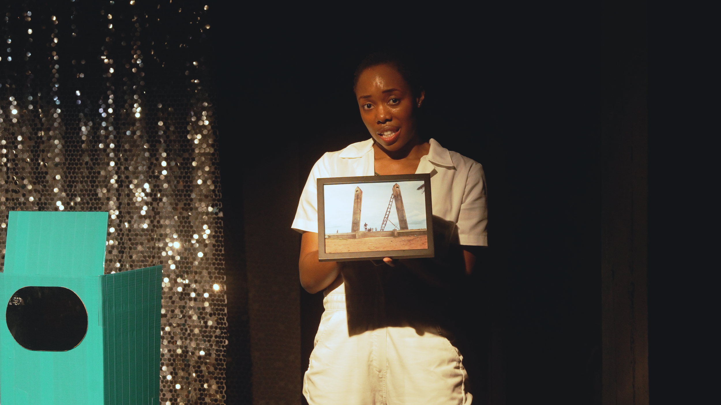 A woman with short curly hair and a white shirt holds a framed photograph of a bridge under construction. The background features a black sequined curtain and partial teal and black props.