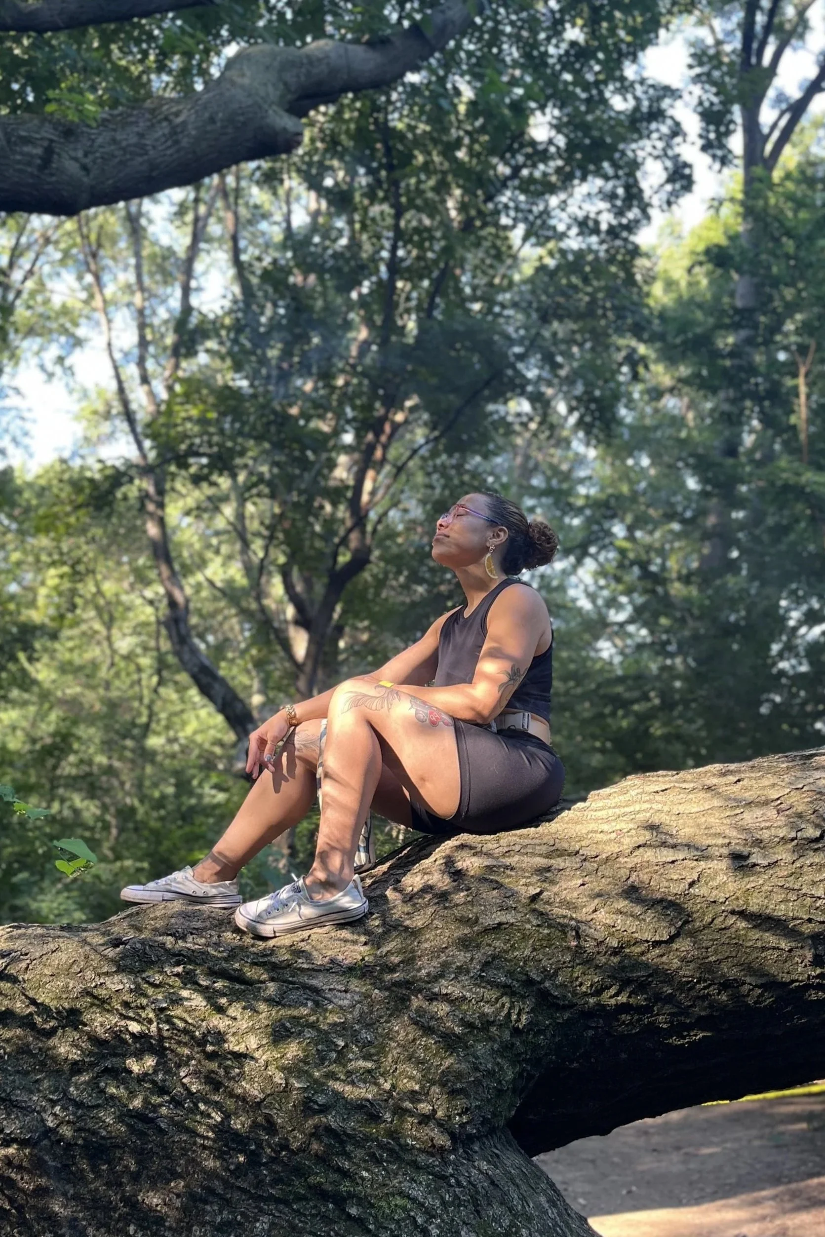 A woman with tattoos, wearing a black sleeveless top, shorts, sneakers, and glasses, sitting on a large tree branch in a forest with green leaves and sunlight filtering through.
