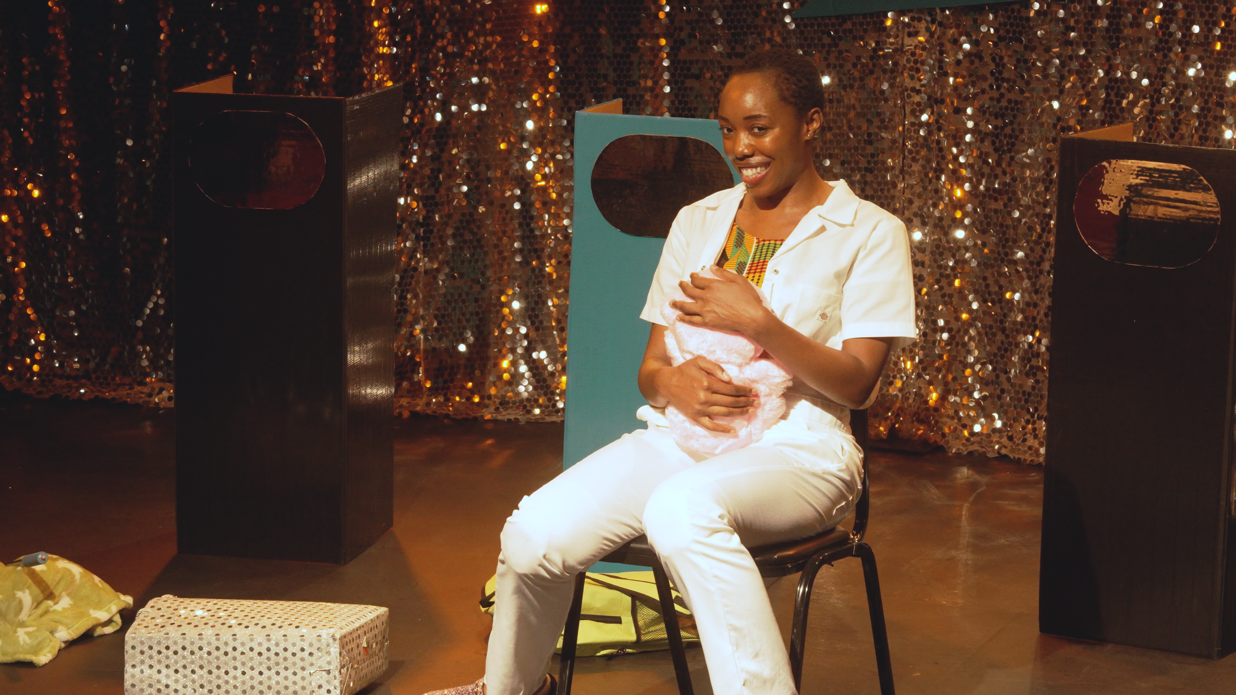 A woman sitting on a chair, holding a pink plush toy, smiling, with a sparkly, sequin-covered backdrop behind her and black speakers on either side.