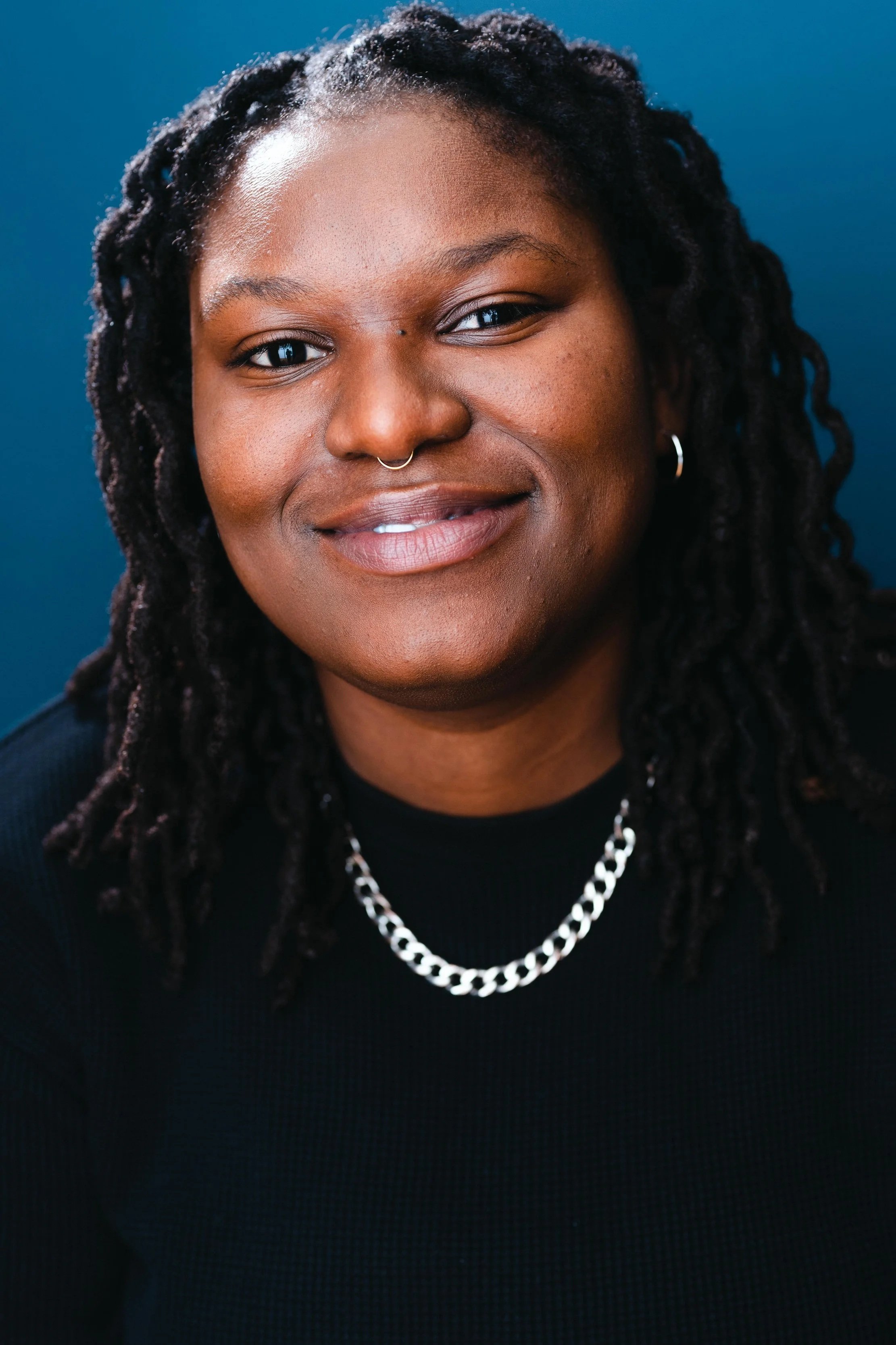 Close-up of a smiling African American woman with dreadlocks, wearing a black top, a silver chain necklace, earrings, and a septum piercing, against a blue background.
