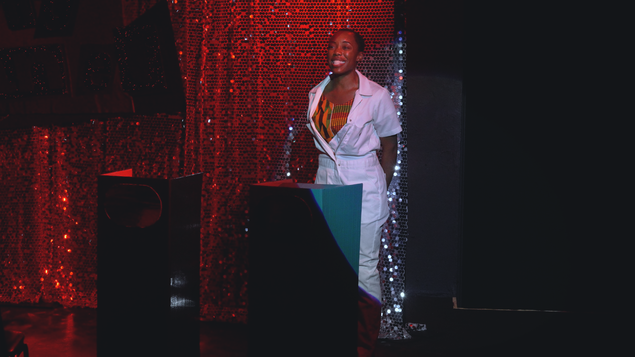 A woman smiling with closed eyes, wearing a white jumpsuit over a colorful, patterned shirt, standing in front of a red, sequin-covered backdrop with some black and white objects in the foreground.