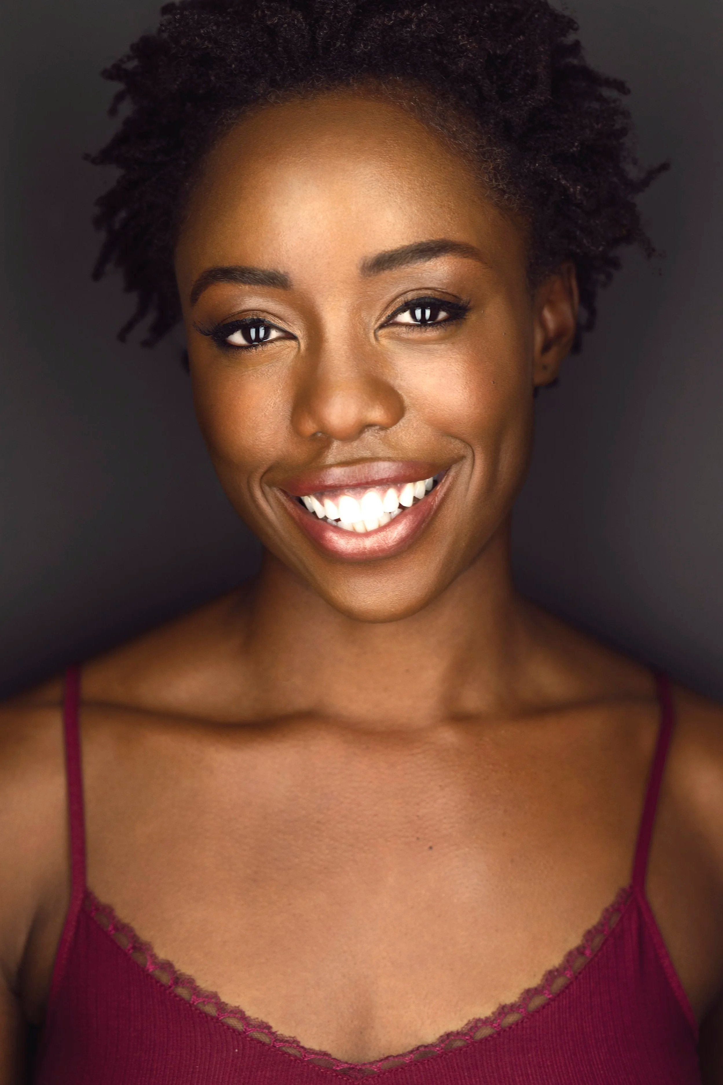Close-up of a smiling woman with curly short hair wearing a purple top with lace trim, standing against a dark background.