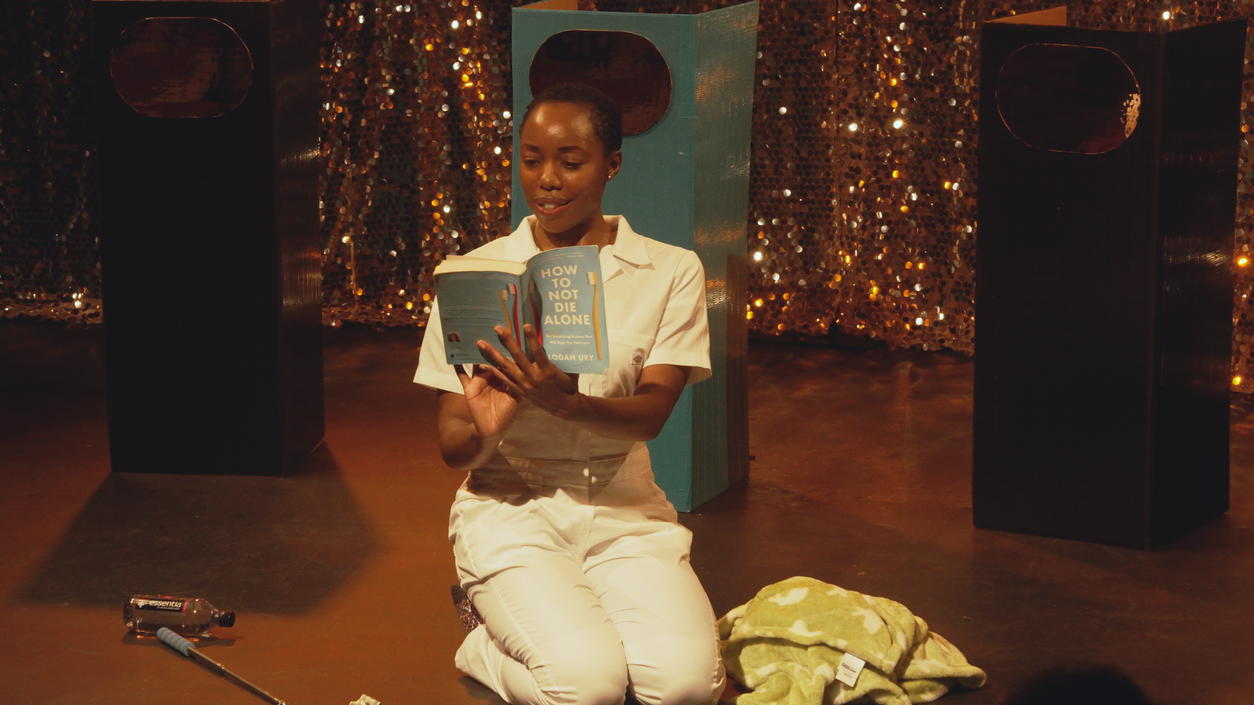 A woman with short hair sitting on the floor, reading a book titled "How to Not Die Alone" in a room with a gold sequin backdrop. She is wearing a white shirt and white pants, and there are some objects on the floor including a flashlight, a marker, 