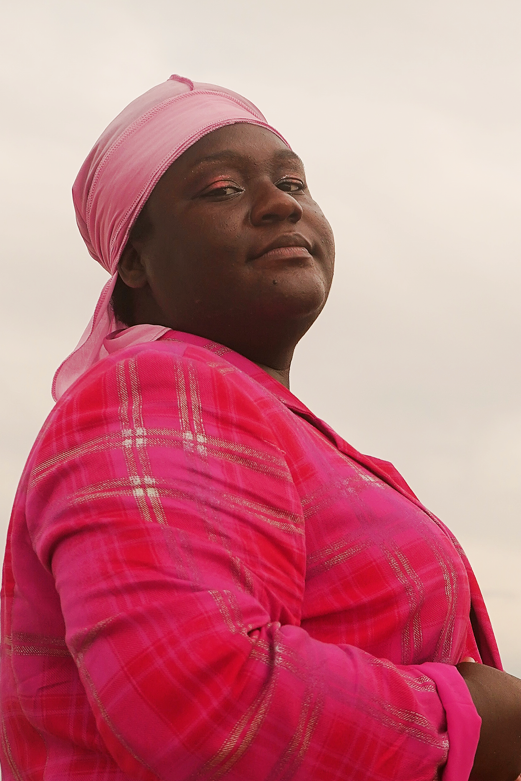 A woman wearing a pink headscarf and matching pink plaid shirt, looking slightly over her shoulder with a neutral expression against a plain background.
