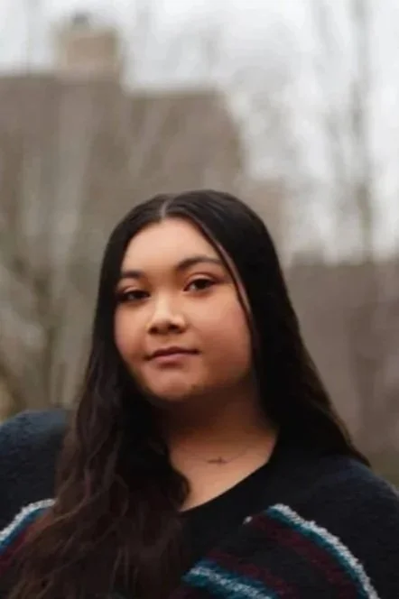 A young girl with long dark hair and tan skin outdoors in front of a blurred natural background.
