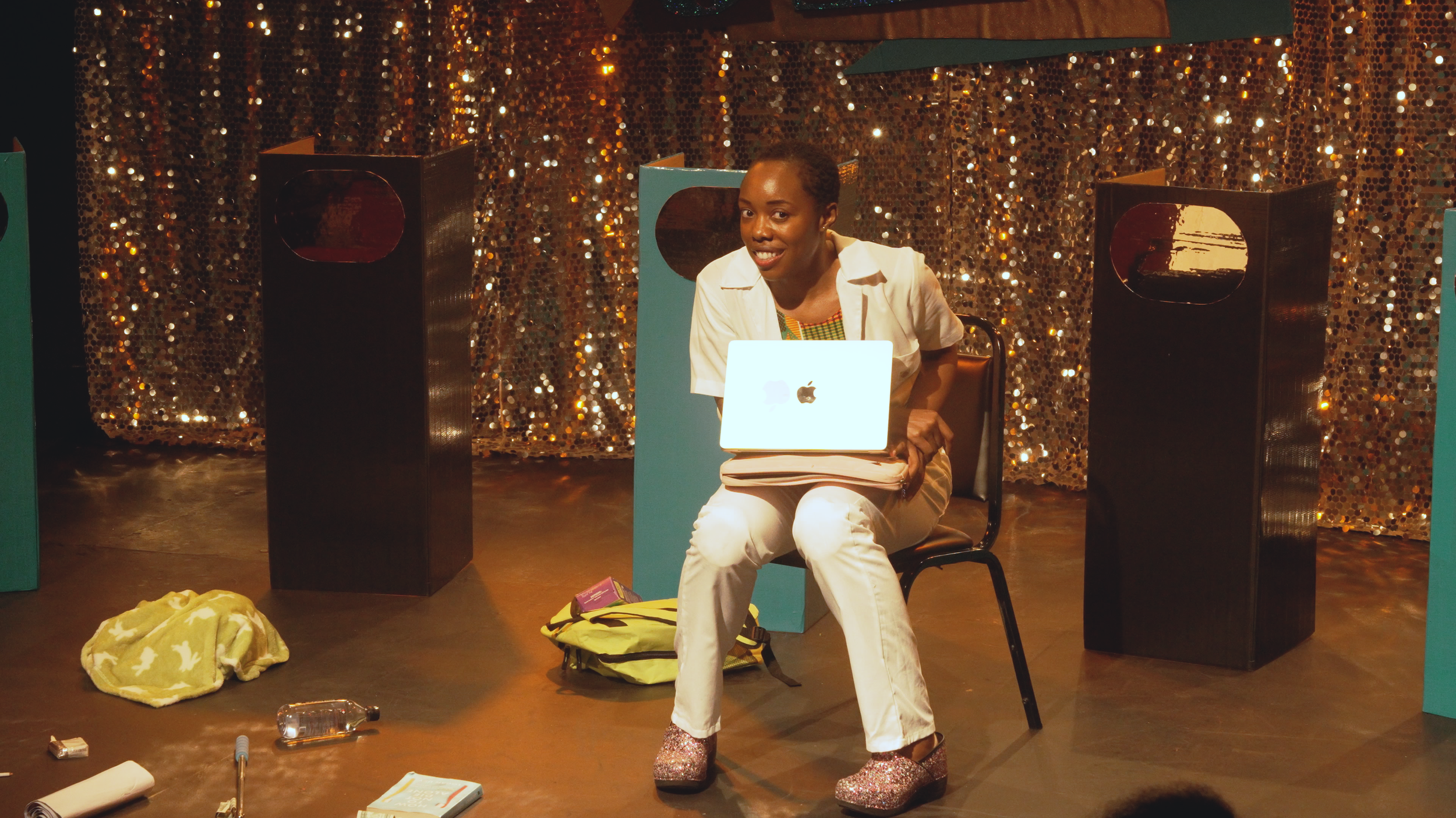 A woman sitting on a chair in front of a gold sequin backdrop, smiling, with a laptop on her lap. There are black display stands around her, and various items and luggage on the floor.