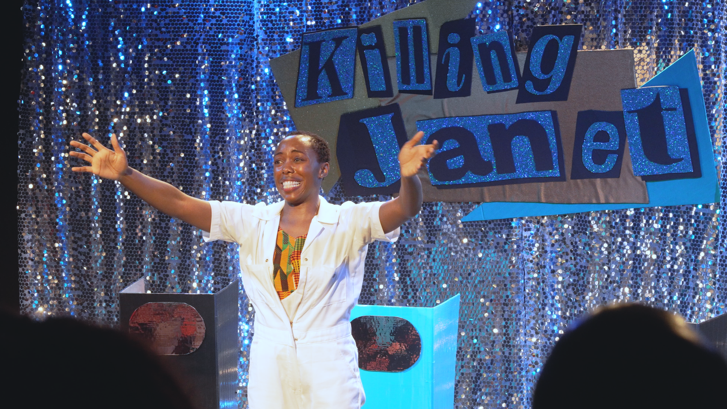 A woman with short hair wearing a white outfit, smiling and gesturing with her arms outstretched on stage with a sparkly blue and silver backdrop. A sign that reads "Killing Jealousy" is behind her.