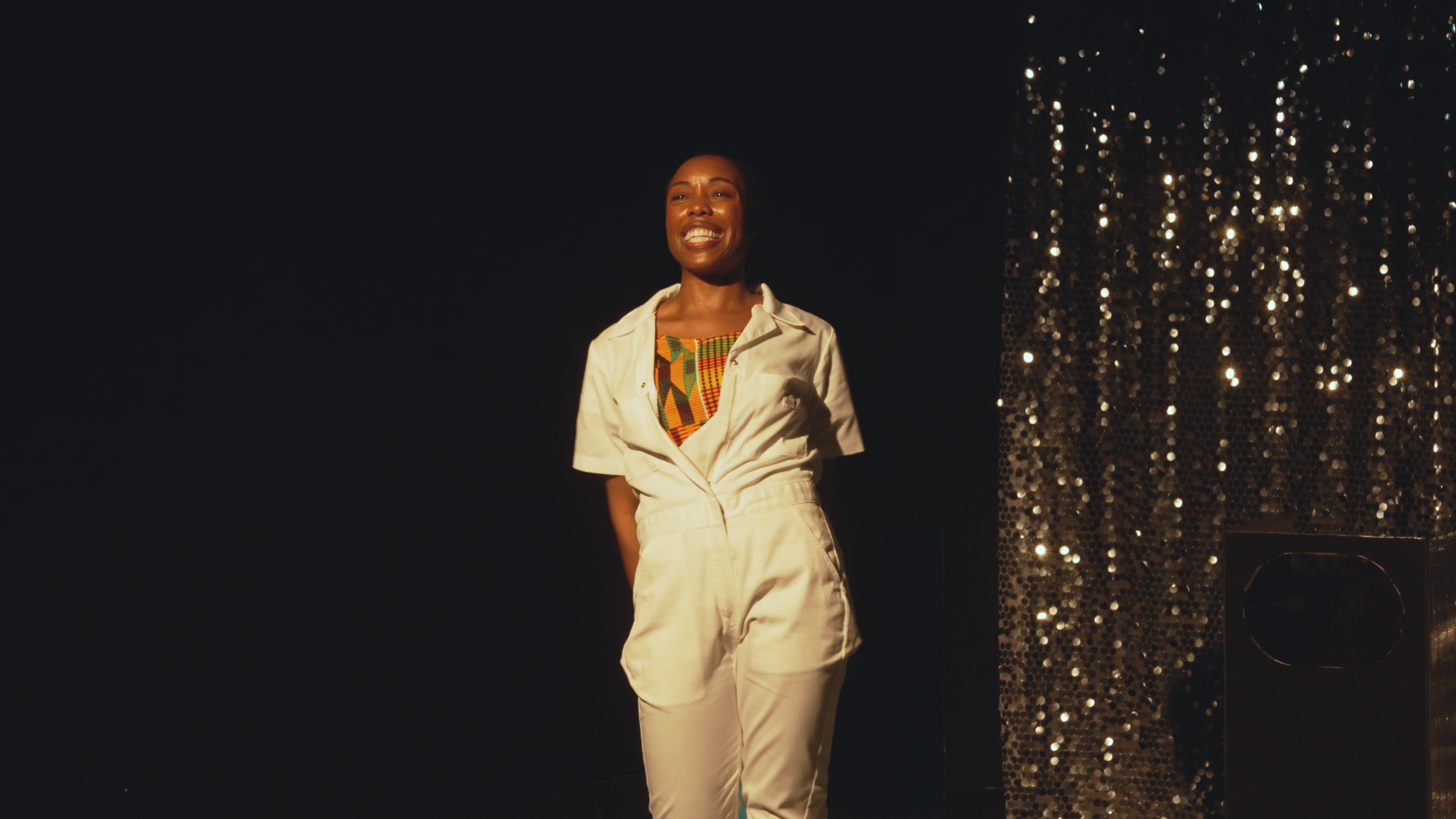 A woman smiling and standing on stage against a black background with a glittery curtain to her right, wearing a colorful patterned top under a white jumpsuit.