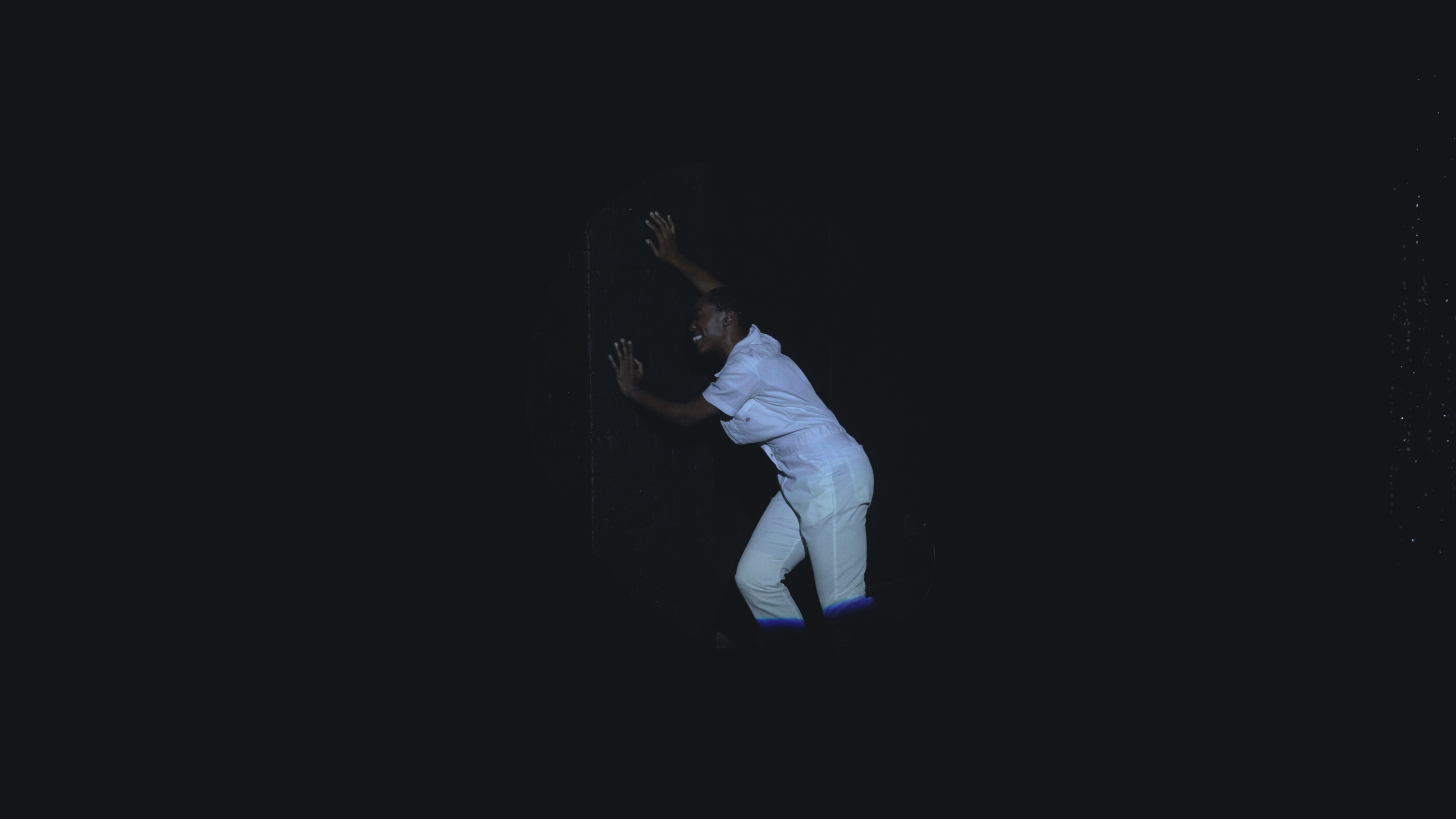 A person dressed in white pants and a white shirt smiling and leaning against a dark wall, with water dripping down the wall.