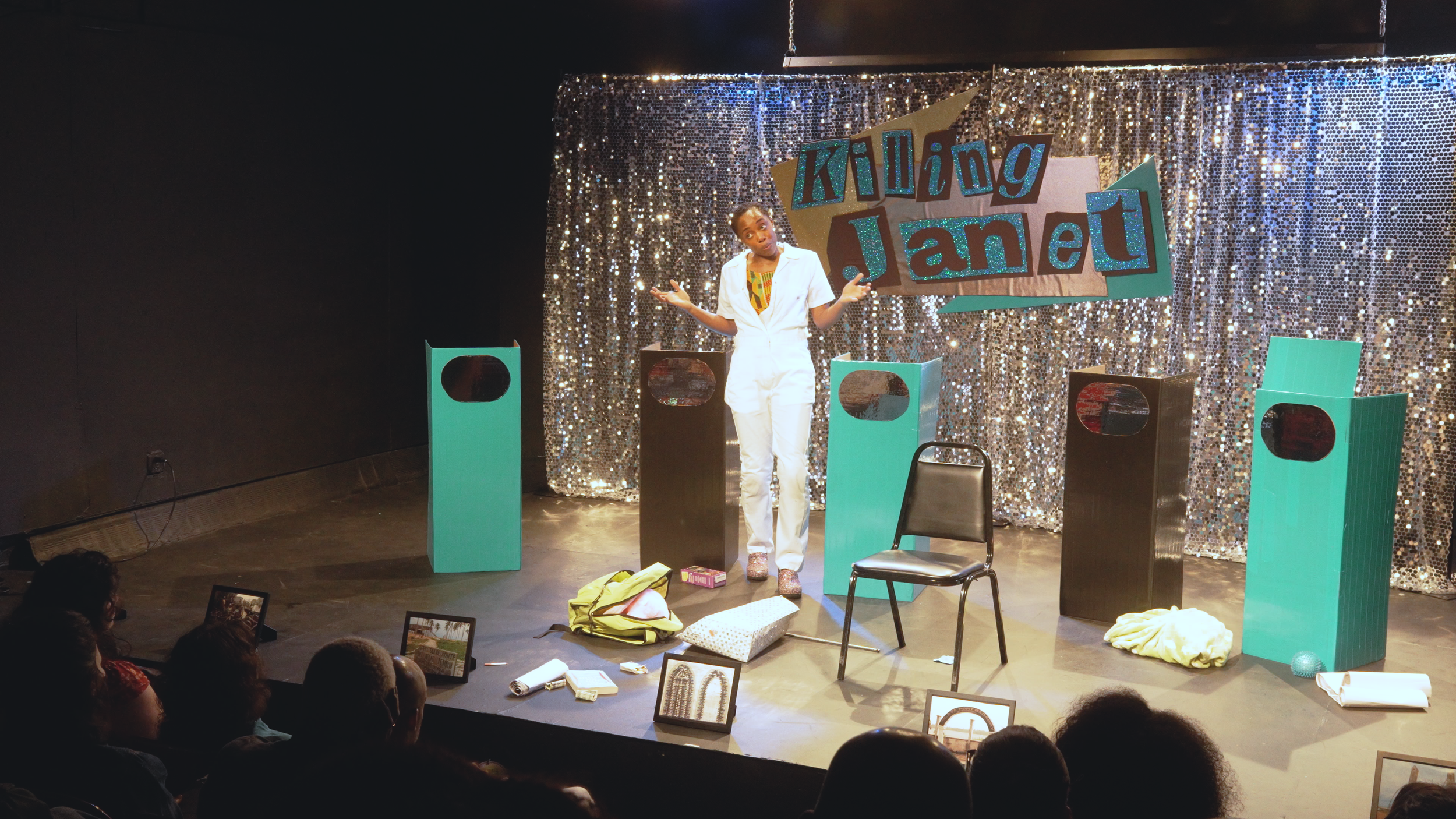 A woman on stage with a sparkly backdrop and a sign that says 'Talking Janel.' She is wearing a white jumpsuit and colorful shoes, gesturing with her hands. There are four turquoise speakers on the stage and various objects, including backpacks and f
