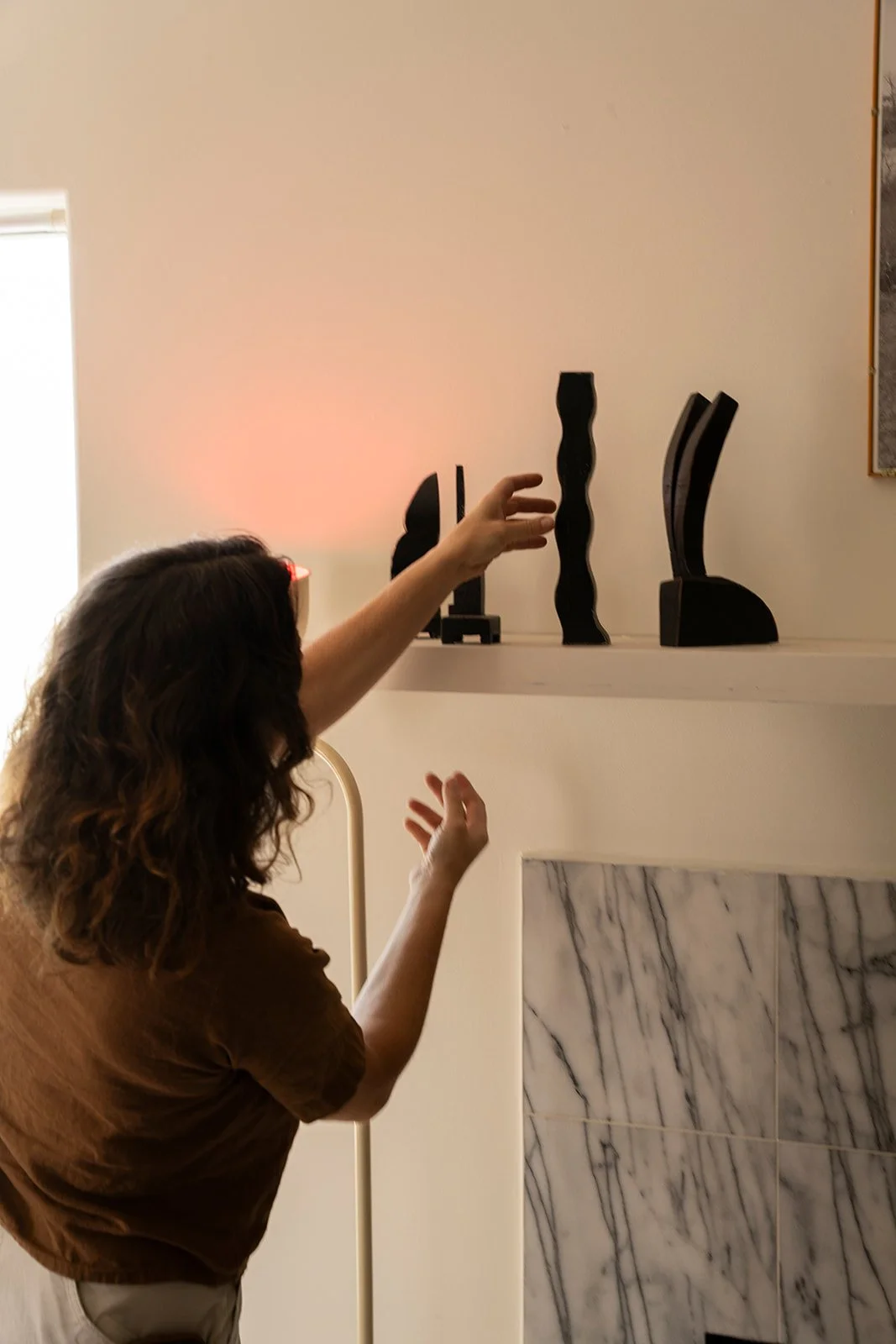 A person with curly hair reaching out toward a display of black sculpture art pieces on a shelf in a room with a marble fireplace.