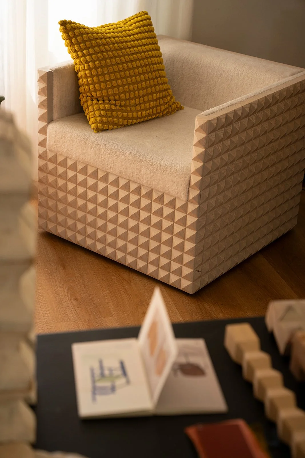 Close-up of a modern beige armchair with geometric textured sides and a mustard yellow blanket with pom-pom details draped over it, in a well-lit room.
