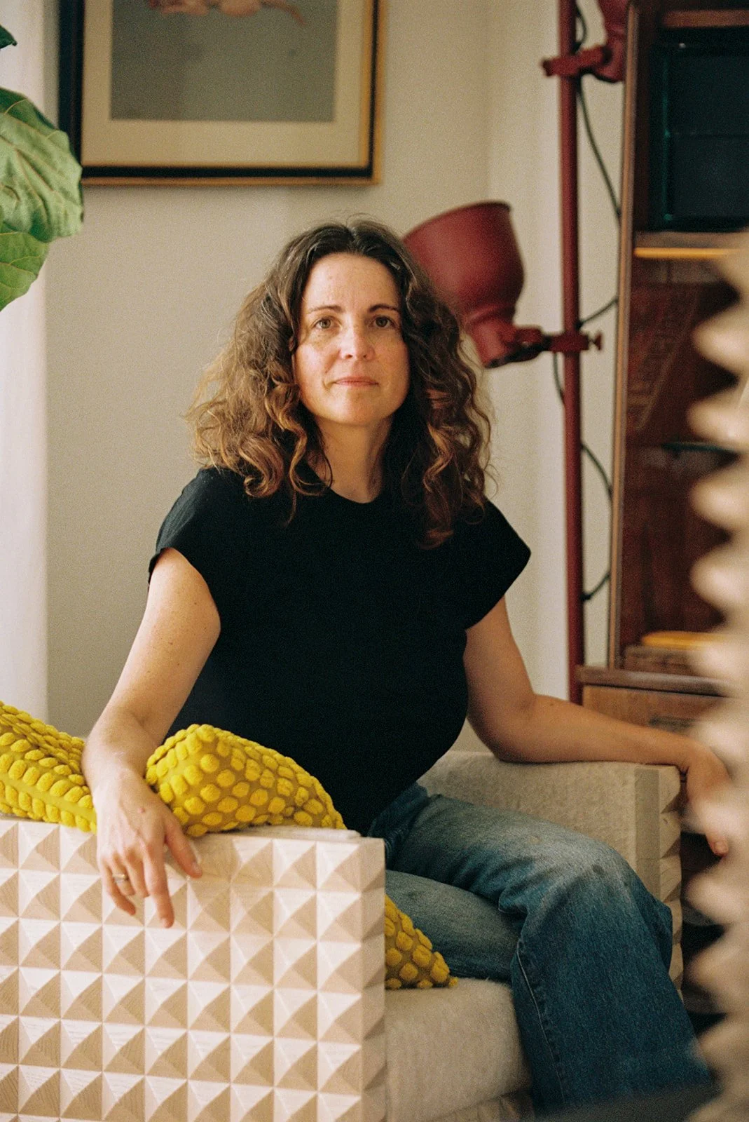A woman with curly brown hair, wearing a black shirt and blue jeans, sitting on a beige couch with a yellow textured blanket, in a cozy living room with artwork, a red lamp, and a bookshelf in the background.