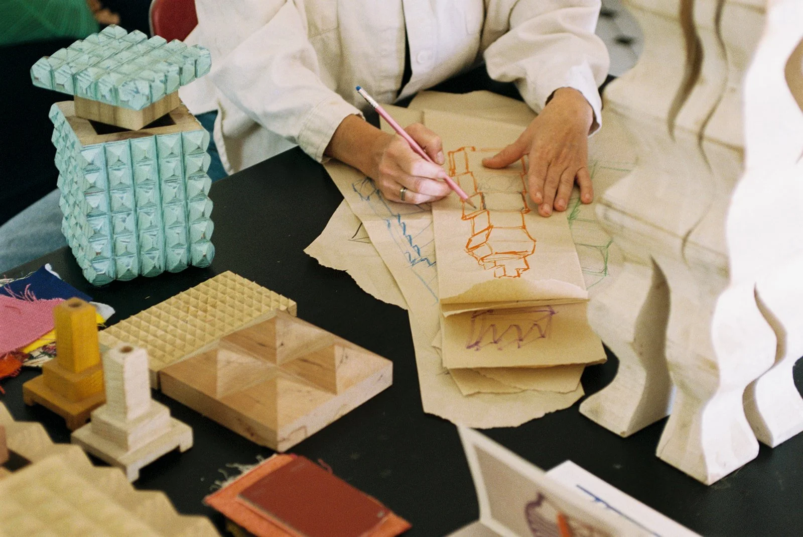 A person working on a craft project at a table with various small wooden and foam models, sketching design plans on paper with a pink pencil.