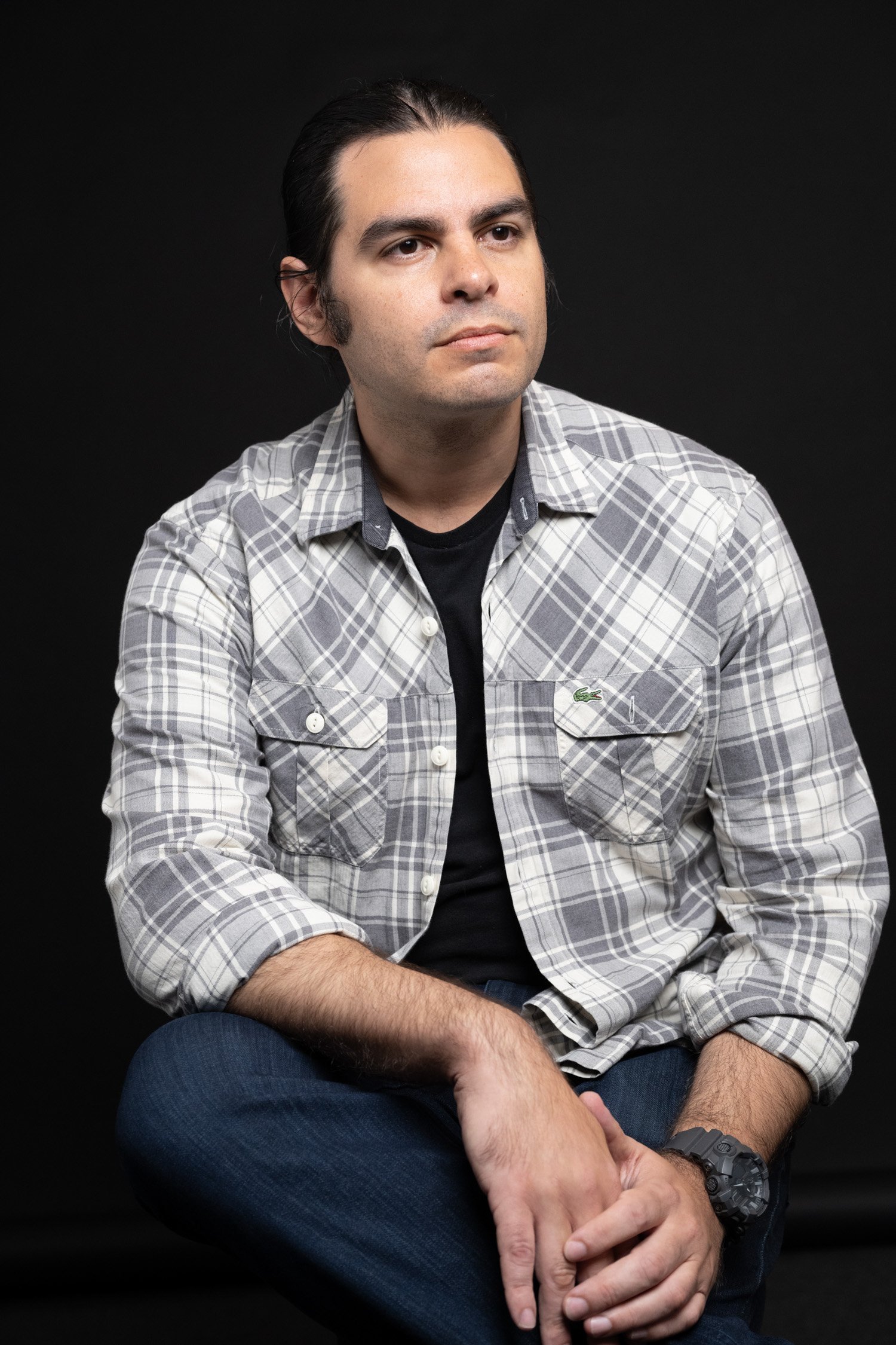 A young man with long dark hair, wearing a gray plaid shirt over a black T-shirt, sitting against a black background, looking to the side with a neutral expression.
